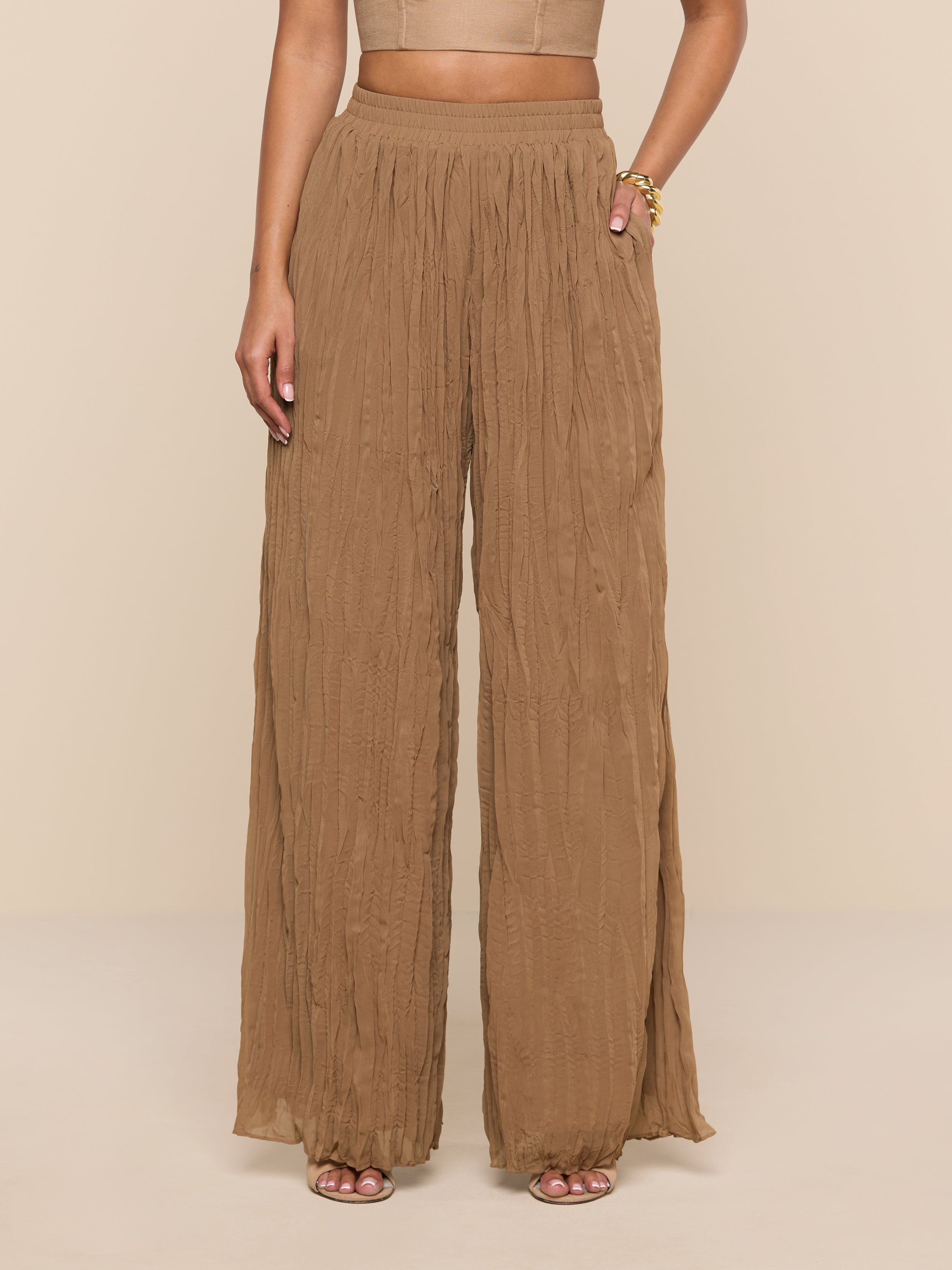 A person wearing the Ethel Pleated Pant, which features a high-waisted, wide-leg silhouette in light brown crinkle chiffon, and sandals stands against a plain white background. Their upper body is partially visible.