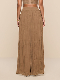 A woman stands facing away, wearing the Ethel Pleated Pant—a loose, high-waisted style in light brown crinkle chiffon—and a sleeveless cropped top against a plain white background.
