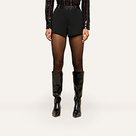 Wearing a black sequin long-sleeve top, sheer black tights, and shiny black knee-high boots, the person showcases the Colt Tuxedo Short—black high-waisted shorts crafted from suiting fabric—while standing against a plain light background.