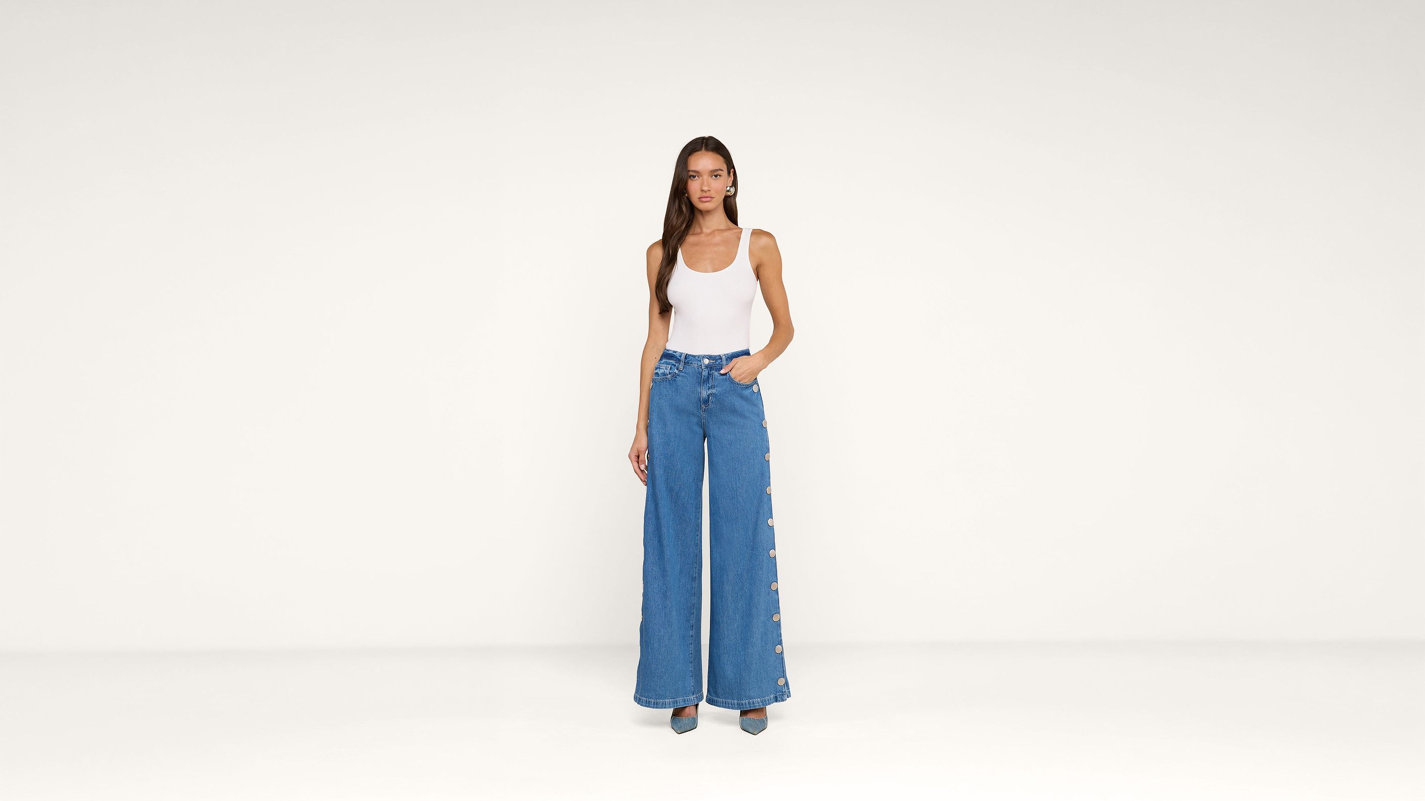 A woman stands against a plain white background, wearing a white sleeveless top and high-waisted wide-leg blue jeans with buttons down the sides. She has long hair and one hand in her pocket.