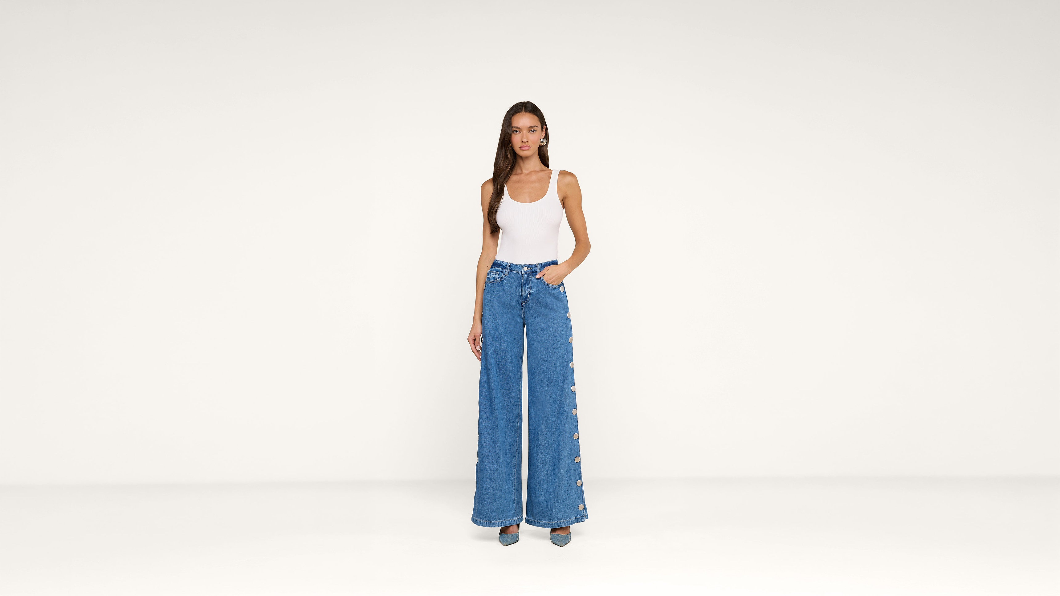 A woman stands against a plain white background, wearing a white sleeveless top and high-waisted wide-leg blue jeans with buttons down the sides. She has long hair and one hand in her pocket.