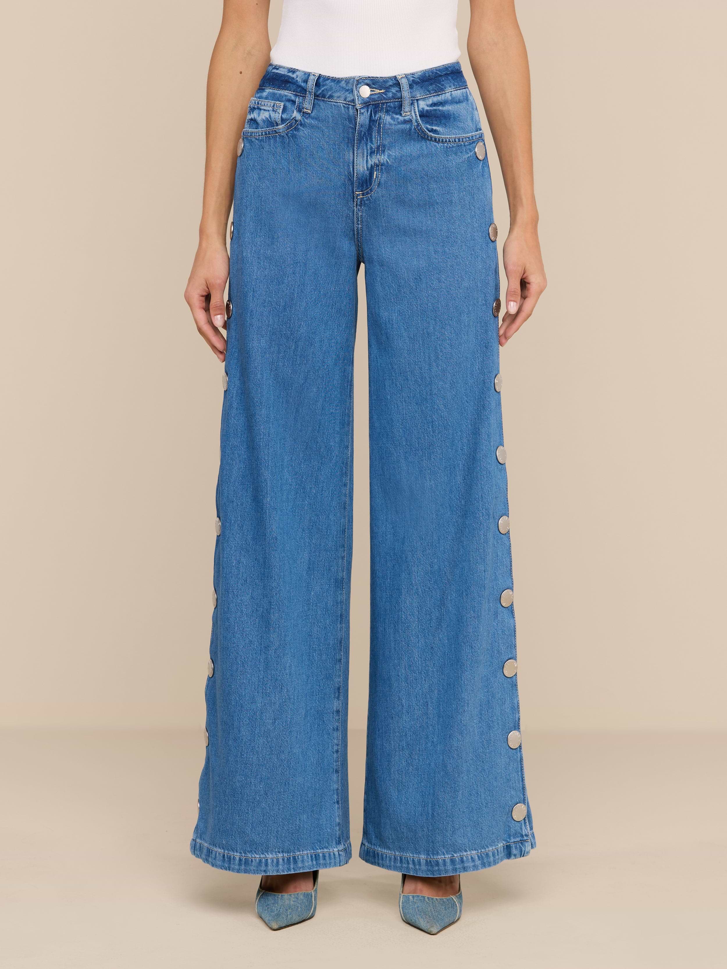 A person wears the Jennings Snap Wide-Leg Jean, featuring high-waisted, wide-leg styling in featherweight denim with large buttons along the outer seams, paired with pointed-toe blue shoes and a white top, standing against a plain white background.