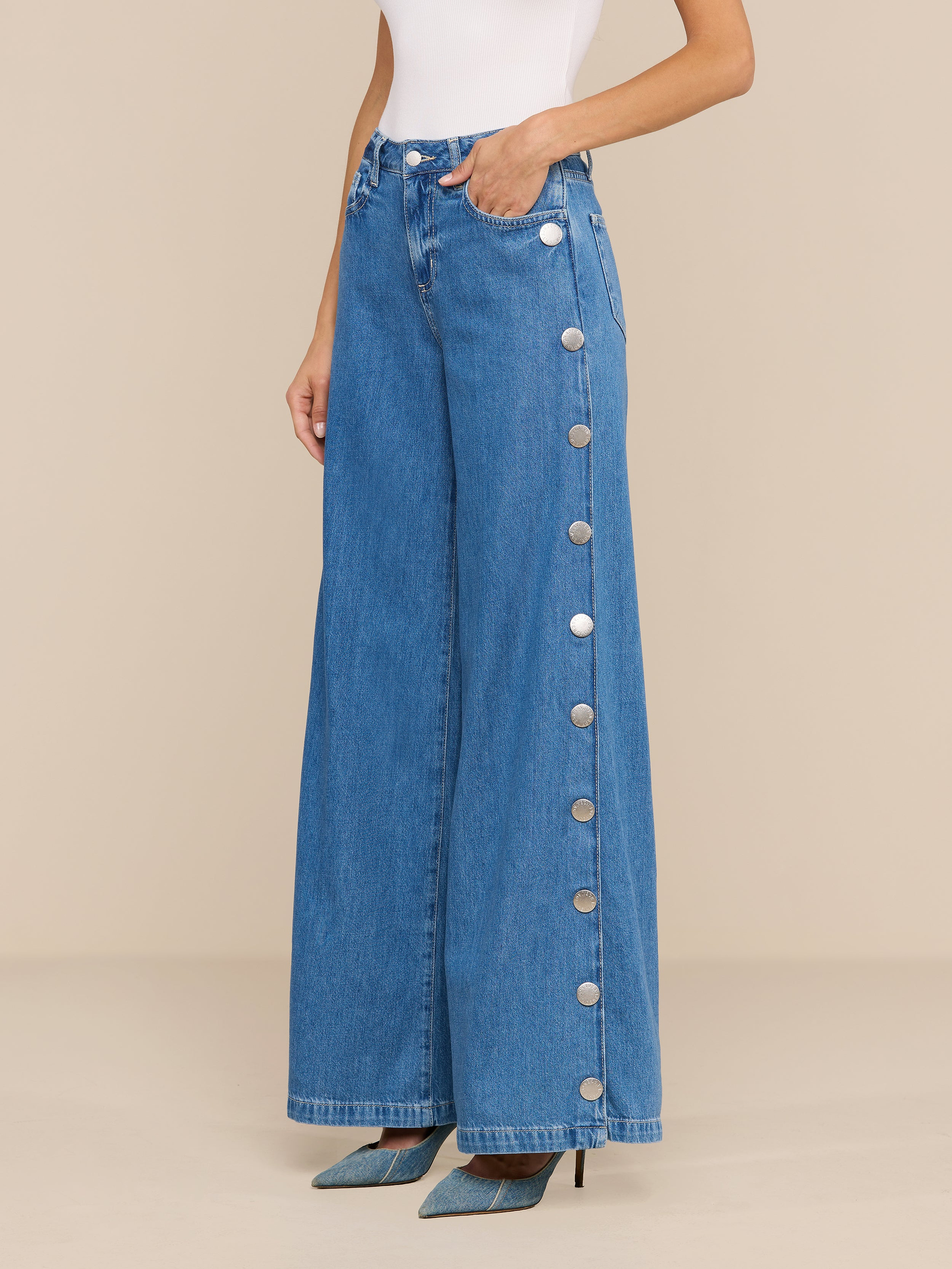 A woman wears the Jennings Snap Wide-Leg Jean, featuring featherweight denim and large silver buttons down the side, styled with a white sleeveless top and pointed-toe high heels, standing against a plain white background.