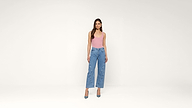 A woman stands against a plain white background, wearing a pink sleeveless top, blue high-waisted jeans, and blue heels. She has long, loose hair and a neutral expression.
