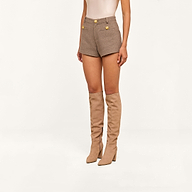 A person wearing the Dionne Tweed Short in a high rise taupe design with gold buttons, paired with tall beige suede boots, standing against a plain white background.