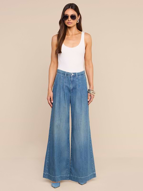 A woman wearing sunglasses, a white tank top, the Jayani Pleated Wide-Leg Jean, and bracelets stands against a plain white background.