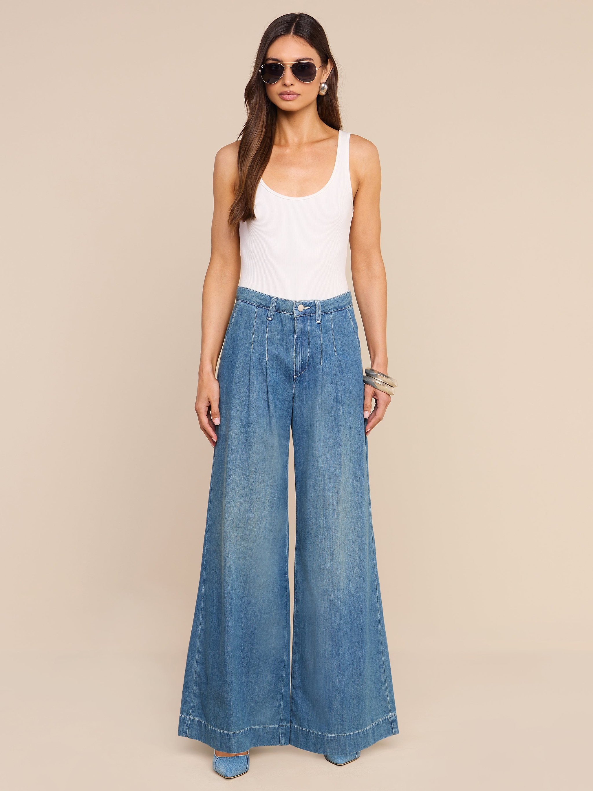A woman wearing sunglasses, a white tank top, the Jayani Pleated Wide-Leg Jean, and bracelets stands against a plain white background.