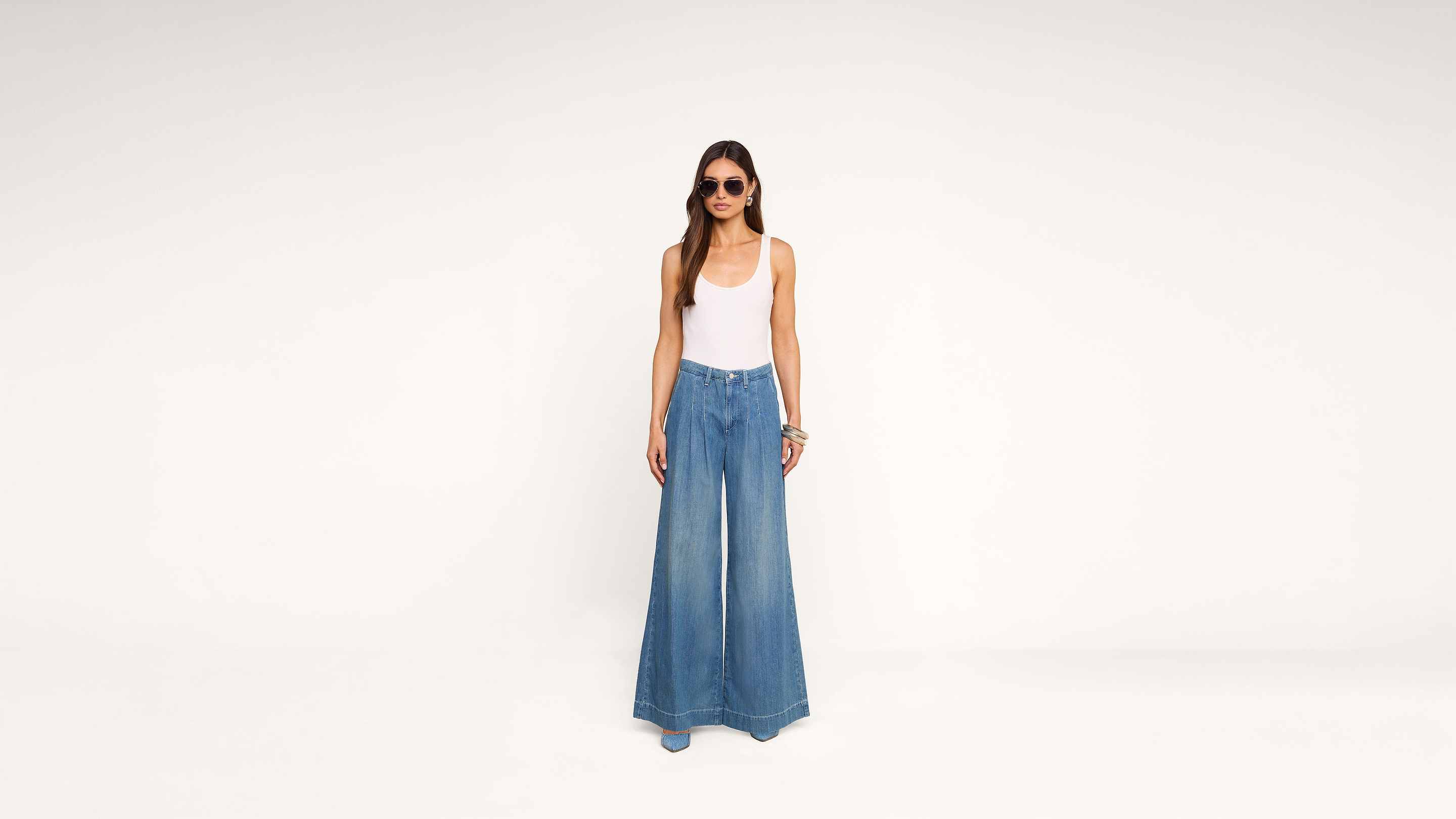 A woman wearing sunglasses, a white tank top, and wide-leg blue jeans stands against a plain white background.