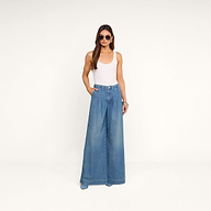A woman wearing a white tank top, the Jayani Pleated Wide-Leg Jean in featherweight denim, sunglasses, and bracelets stands with her hands in her pockets against a plain white background.