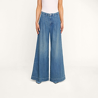 신축성이 없는 데님 소재로 제작된 블루 컬러의 ‘자야니 플리츠 와이드 레그 진’과 흰색 민소매 상의를 입은 이 사람은 베이지색 배경 앞에 서서 양손을 몸 옆에 내려놓고 있습니다. 하반신과 다리만 보입니다.