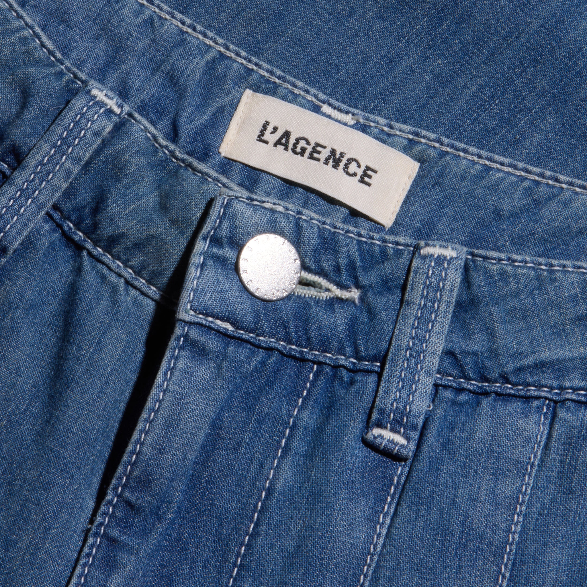 Close-up of the Jayani Pleated Wide-Leg Jean in blue denim, highlighting the waistband, a silver button, belt loop, and a white label with black LAGENCE text. Crafted from featherweight denim, these high-rise jeans combine comfort with timeless style.
