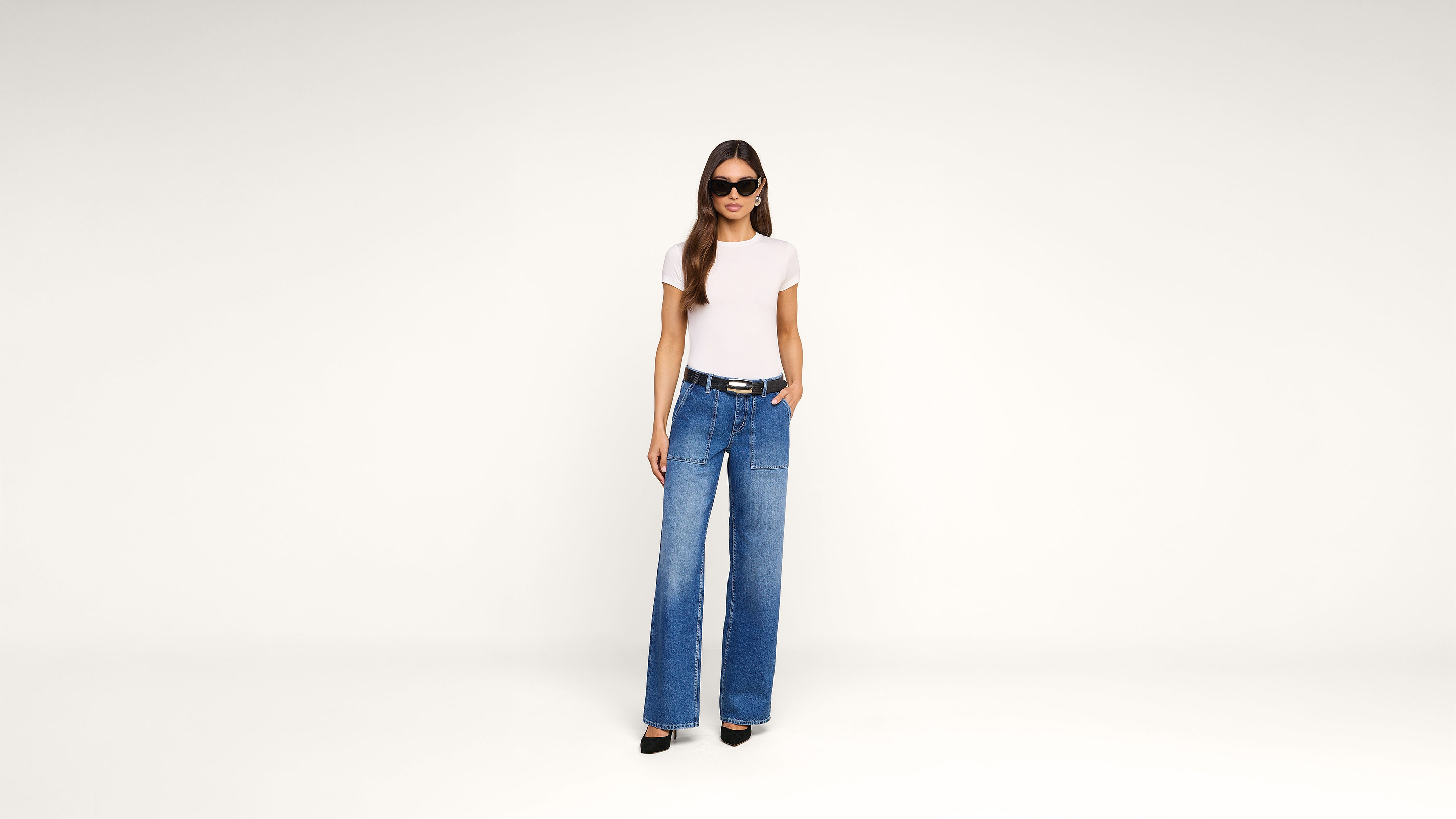 A woman wearing sunglasses, a white t-shirt, and blue wide-leg jeans stands against a plain white background with one hand in her pocket.