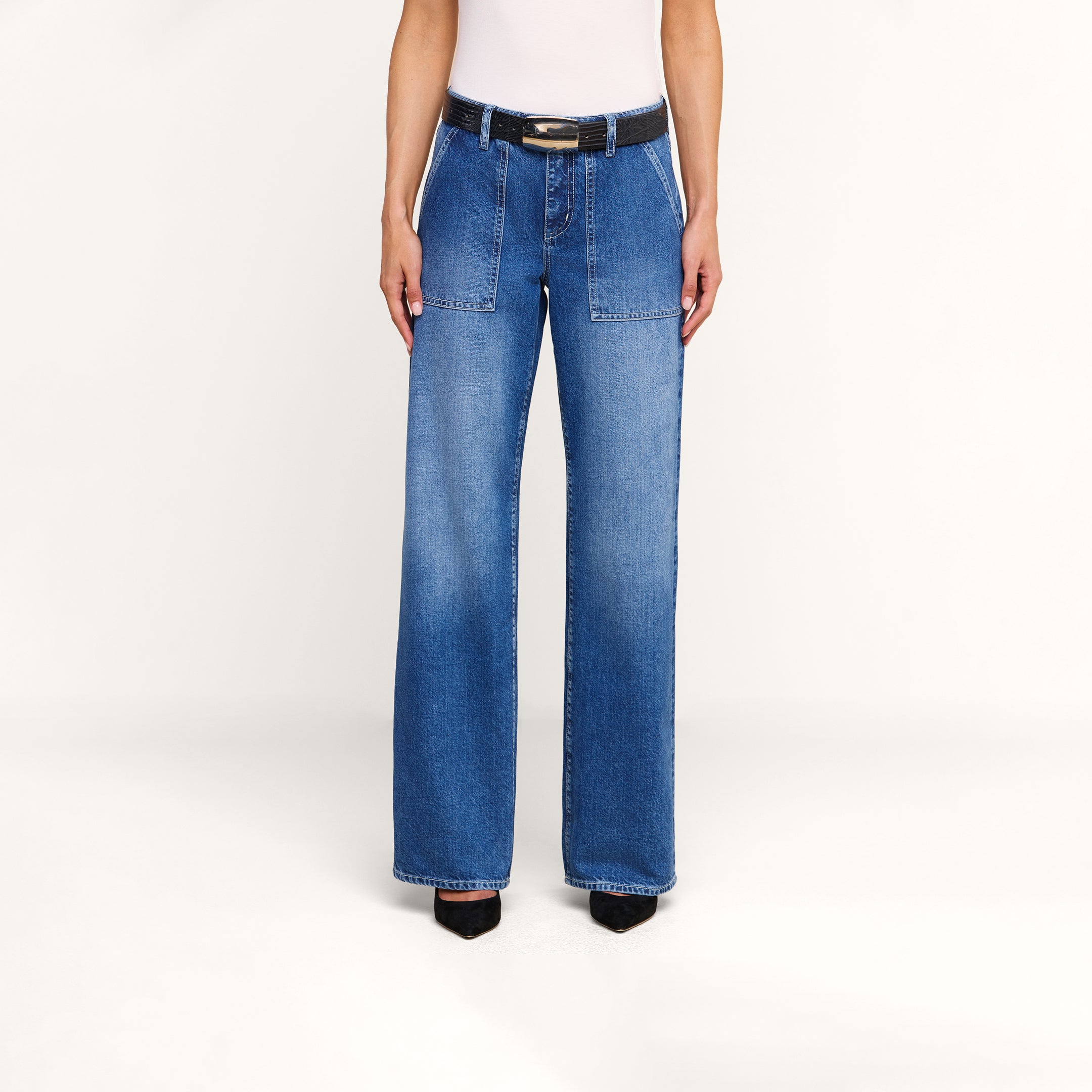 Una persona que lleva los vaqueros «Christel Wide-Leg», un modelo de talle alto y pierna ancha confeccionado en denim 100  algodón con bolsillos de parche delanteros, combinados con un cinturón negro y zapatos negros de punta. La imagen muestra únicamente el torso y las piernas sobre un fondo beige liso.