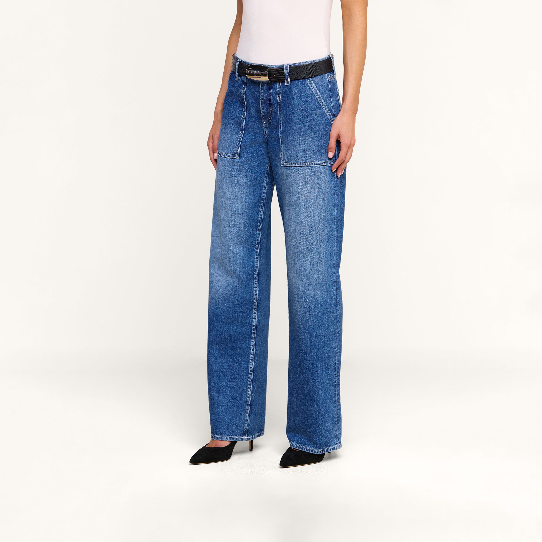 Alguien lleva puestos los vaqueros «Christel Wide-Leg», unos vaqueros azules de cintura alta y corte ancho, confeccionados en denim 100  algodón y con bolsillos de parche delanteros. El conjunto incluye un cinturón negro, unos zapatos de tacón negros de punta afilada y un top blanco sin mangas. El fondo es de color beige liso.