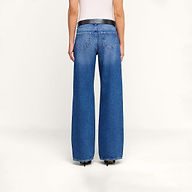 Se muestra a una persona de espaldas sobre un fondo liso de color beige, con unos vaqueros «Christel Wide-Leg» de cintura alta y pierna ancha, confeccionados en denim azul 100  algodón, combinados con un cinturón negro y un top blanco.
