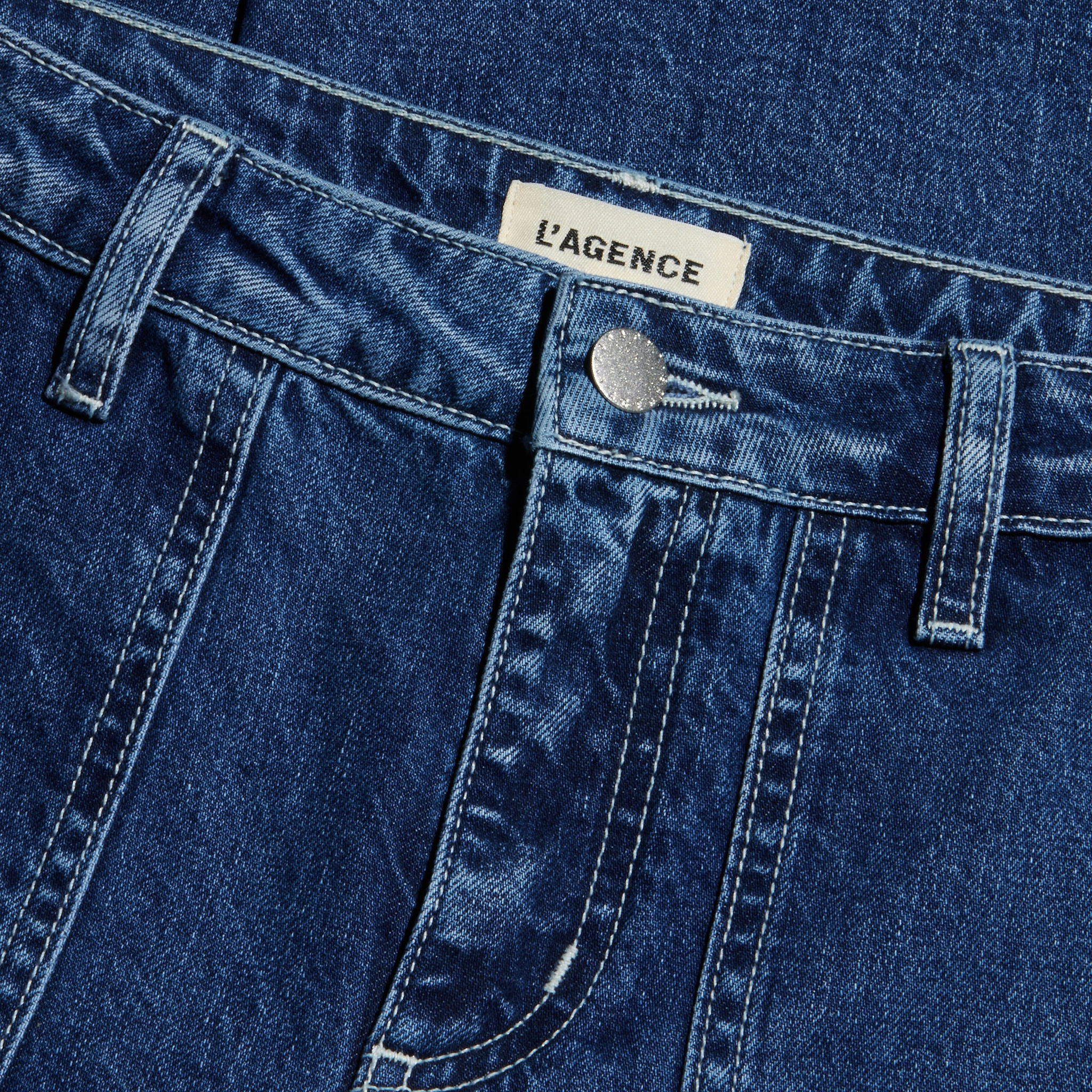 Close-up of blue denim Christel Wide-Leg Jean featuring a metal button, belt loops, visible stitching, wide-leg cut, and a white label with the brand name LAGENCE sewn on the waistband.