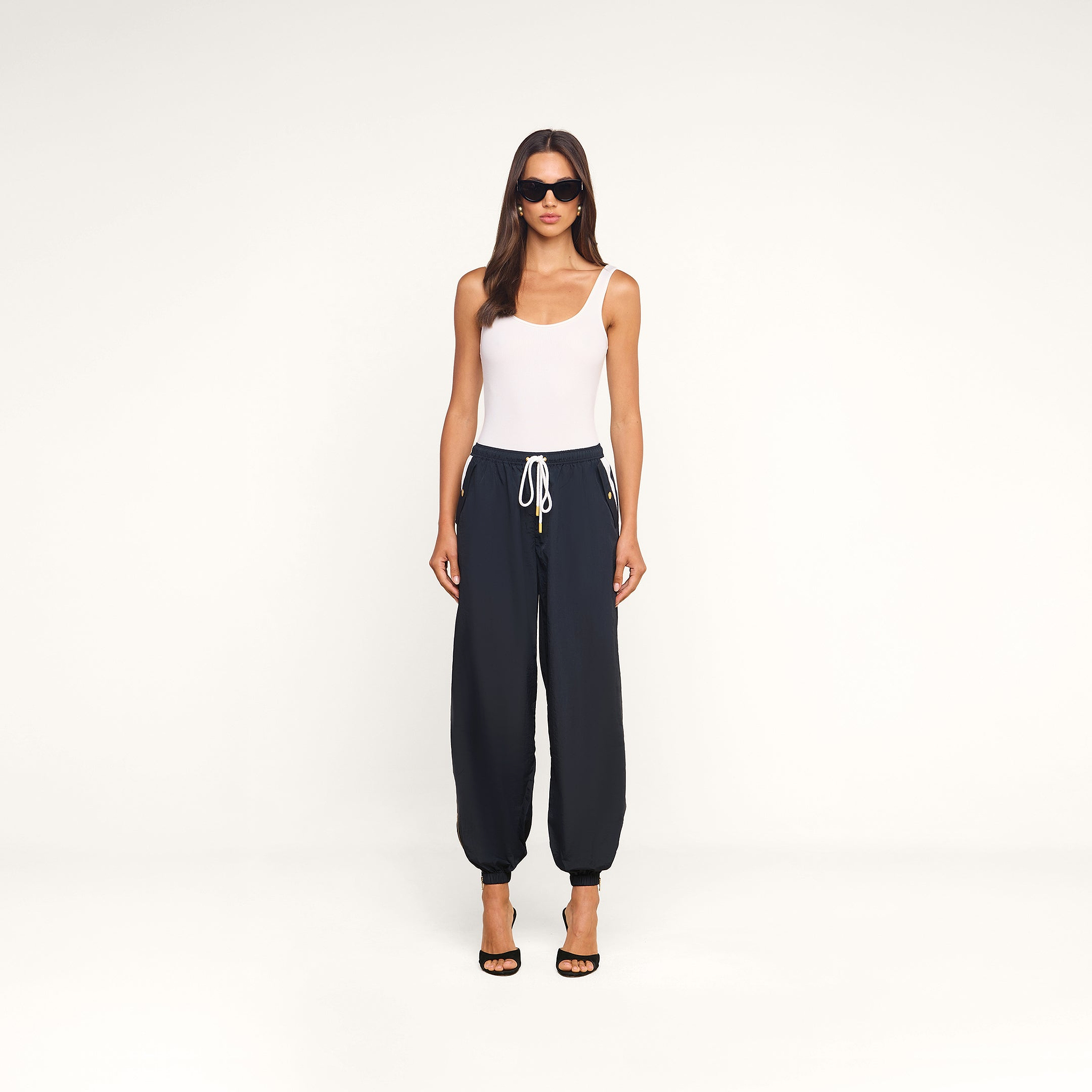 A woman wearing sunglasses, a white sleeveless top, and the Mariko Jogger black lightweight nylon pants stands facing forward against a plain white background. She completes her look with black high-heeled sandals.