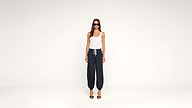 A woman stands against a plain white background, wearing sunglasses, a white tank top, dark drawstring pants, and black high-heeled sandals.
