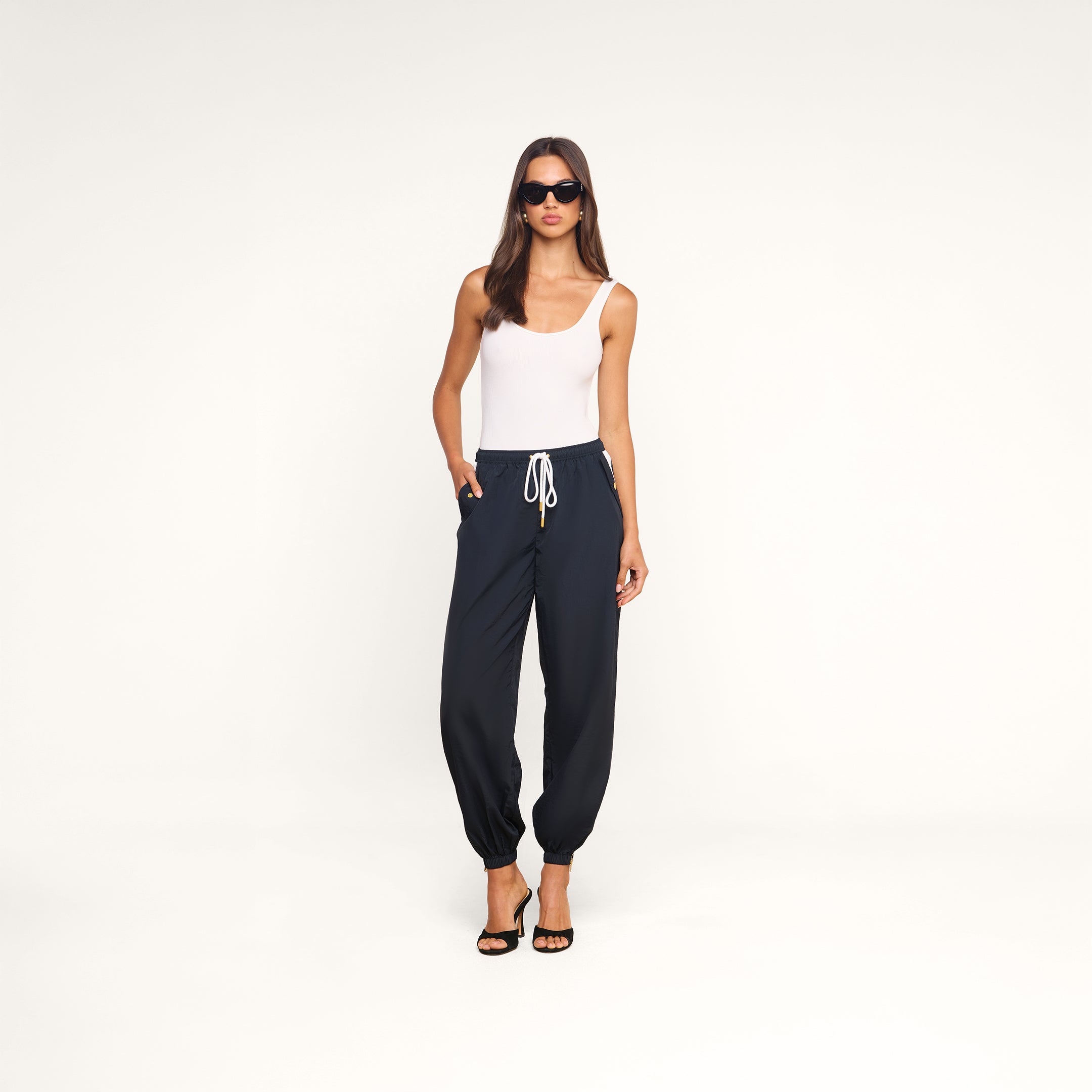 A woman in sunglasses wears a white tank top, the navy blue Mariko Jogger made from lightweight, water-repellent nylon, and black heeled sandals while standing against a plain white background.