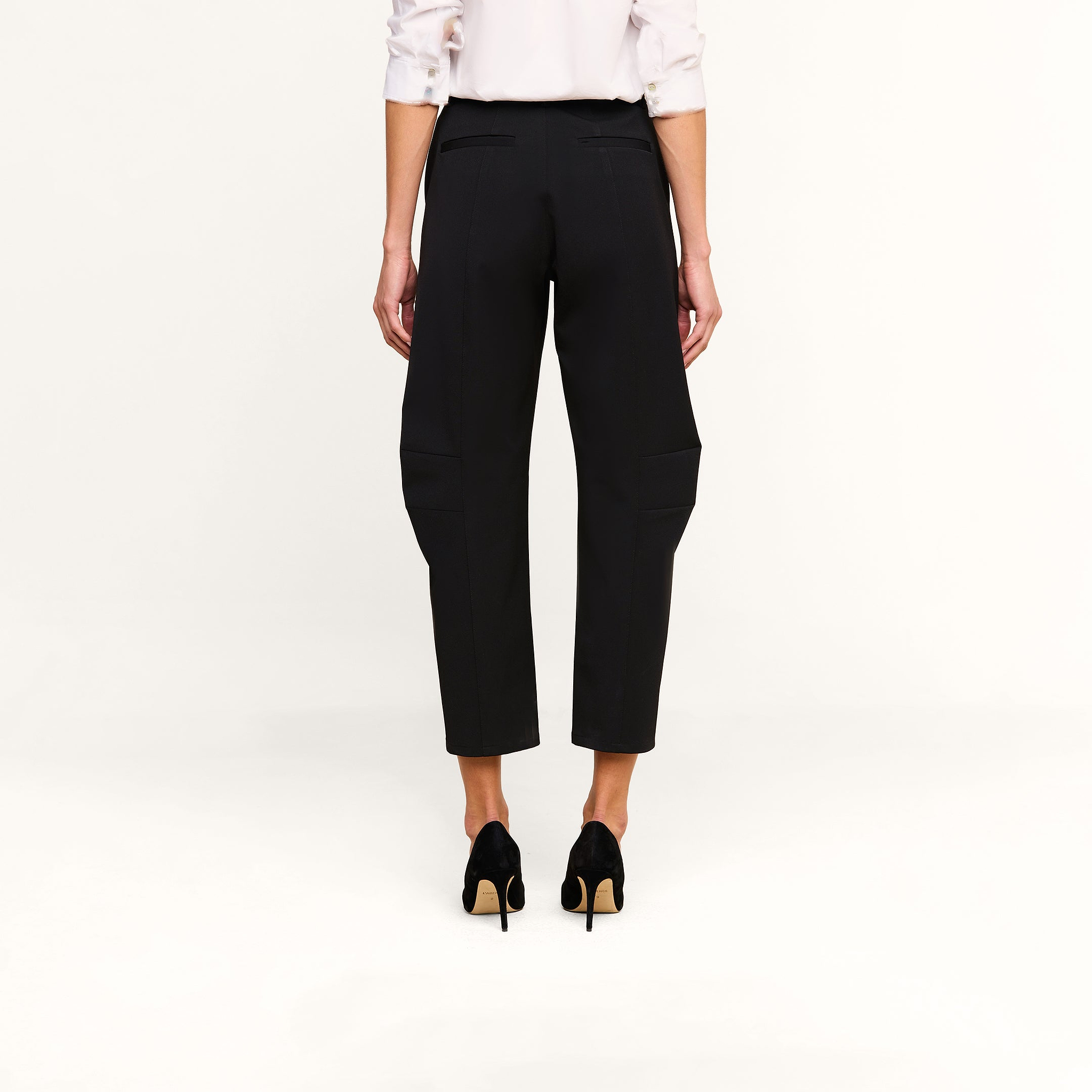 A person stands facing away against a plain white background, wearing the Colby Barrel-Leg Trouser in black, paired with a white long-sleeve shirt with rolled-up sleeves and black high-heeled shoes.