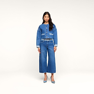 Una mujer posa de frente, vestida con una chaqueta vaquera corta y los vaqueros «Bronte Cropped Wide-Leg», que destacan por su cintura alta y su corte corto de pierna ancha. Completa el look con unos zapatos de tacón azules sobre un fondo beige liso.