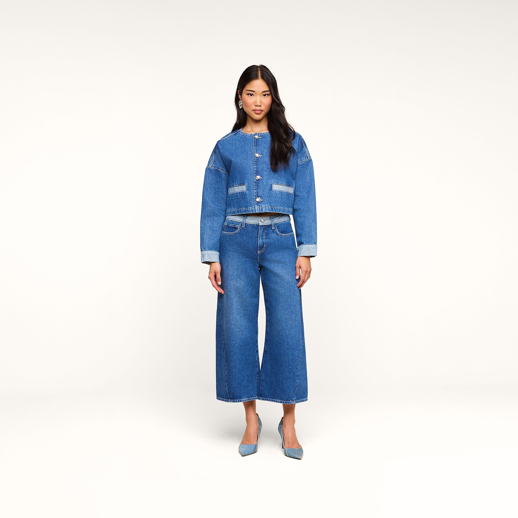 Una mujer posa de frente, vestida con una chaqueta vaquera corta y los vaqueros «Bronte Cropped Wide-Leg», que destacan por su cintura alta y su corte corto de pierna ancha. Completa el look con unos zapatos de tacón azules sobre un fondo beige liso.