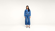 A woman stands against a plain white background, wearing a blue denim jacket, matching wide-leg jeans, a belt, and blue heels.
