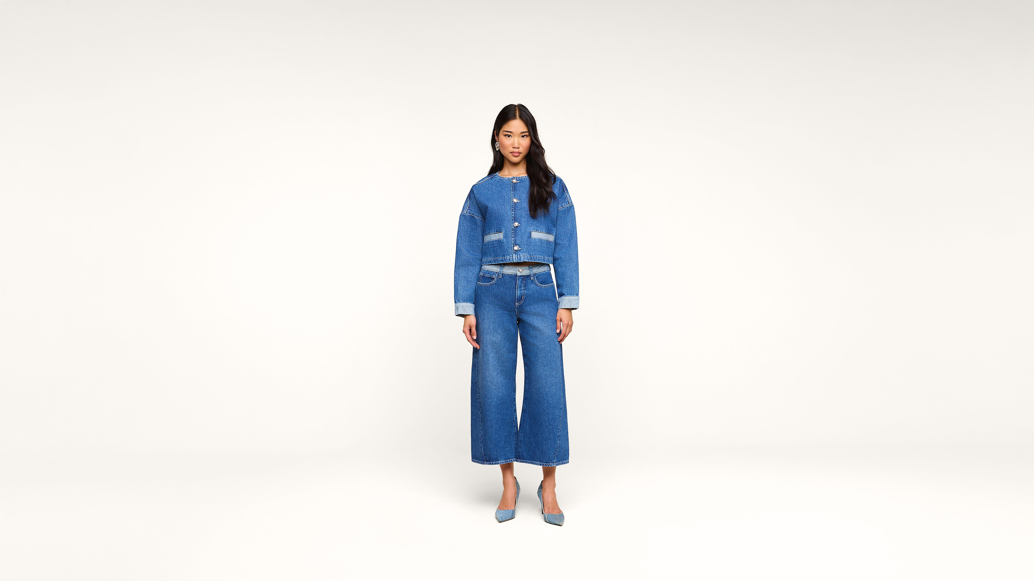 A woman stands against a plain white background, wearing a blue denim jacket, matching wide-leg jeans, a belt, and blue heels.
