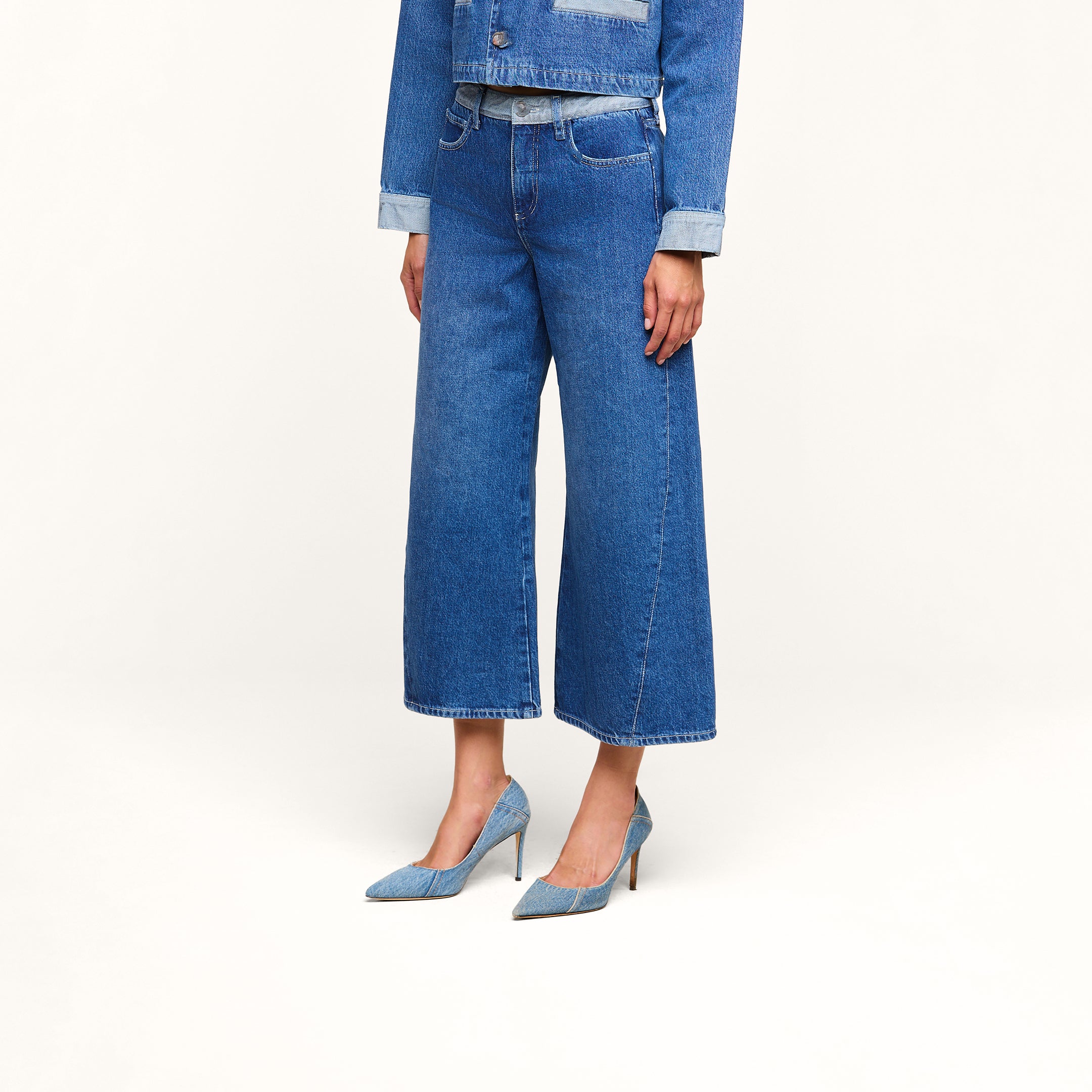 Una persona que lleva unos vaqueros cortos de pierna ancha «Bronte» en color azul, combinados con una chaqueta vaquera a juego y unos zapatos de tacón azul, posa frente a un fondo beige sin que se le vea la cara.