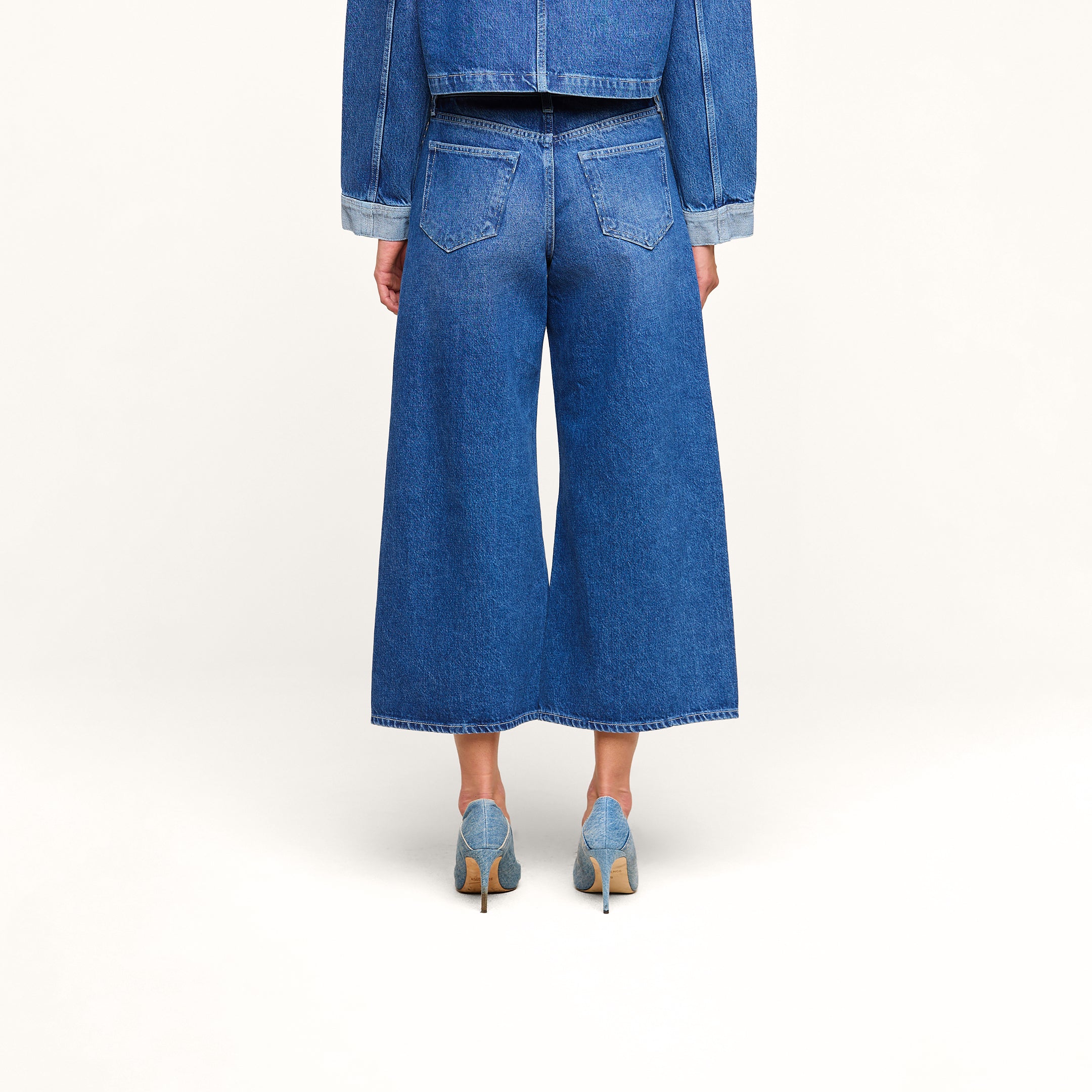 Una modelo luce los vaqueros cortos de pierna ancha «Bronte», de cintura alta, combinados con una chaqueta vaquera y unos zapatos de tacón azul claro; la imagen muestra su espalda sobre un fondo liso de color beige.