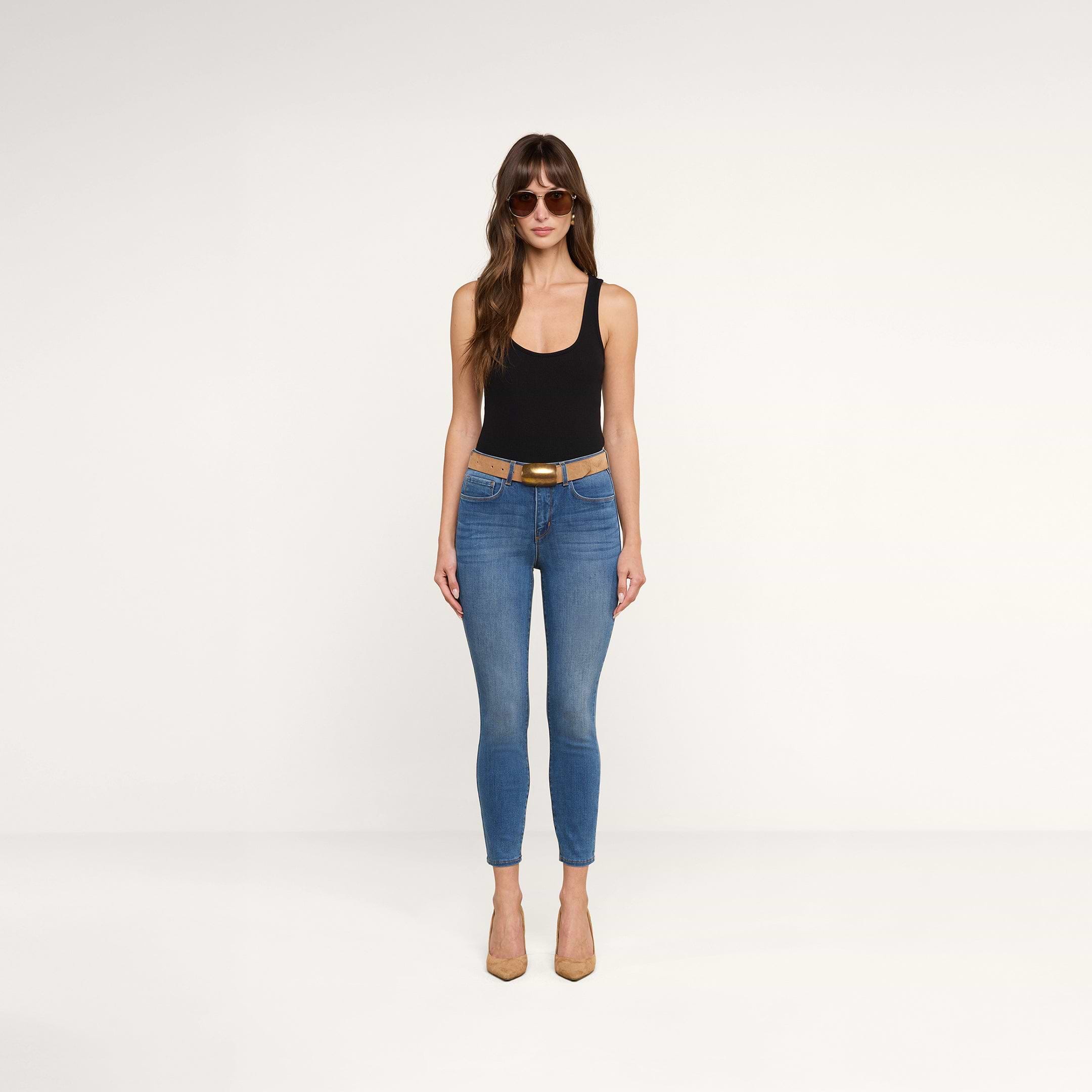 Une femme portant des lunettes de soleil, un haut noir sans manches, un jean skinny court « Margot » au délavage vintage, une ceinture beige clair et des talons beiges clairs se tient de face devant un fond beige uni.