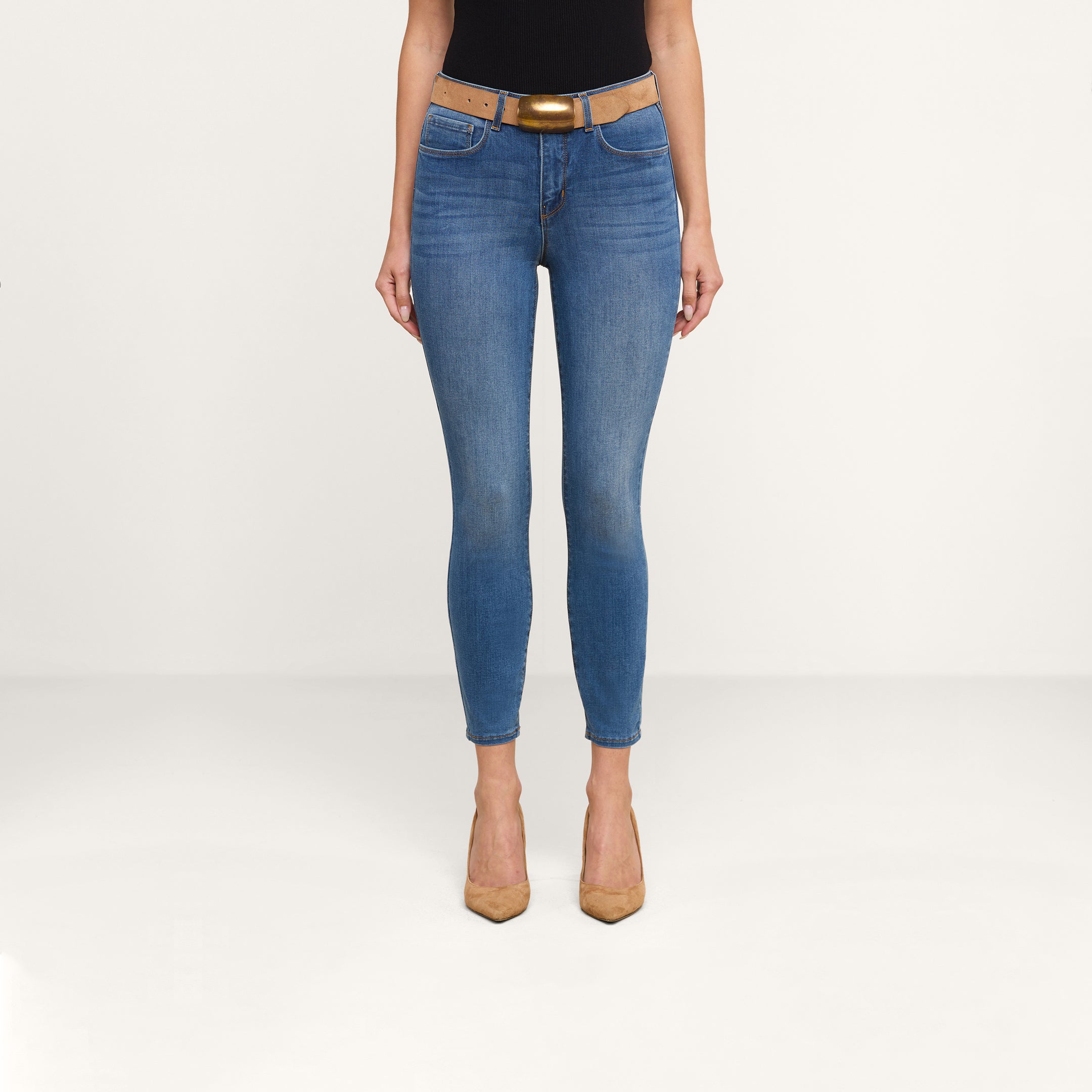 Vêtue dun jean skinny court « Margot » au délavage vintage, dun haut noir, dune ceinture beige clair à boucle dorée et descarpins beiges clairs, la personne se tient debout devant un fond beige uni.