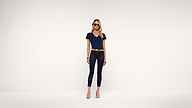 A woman wearing sunglasses, a navy t-shirt, black pants, a yellow belt, and nude heels stands against a plain white background.