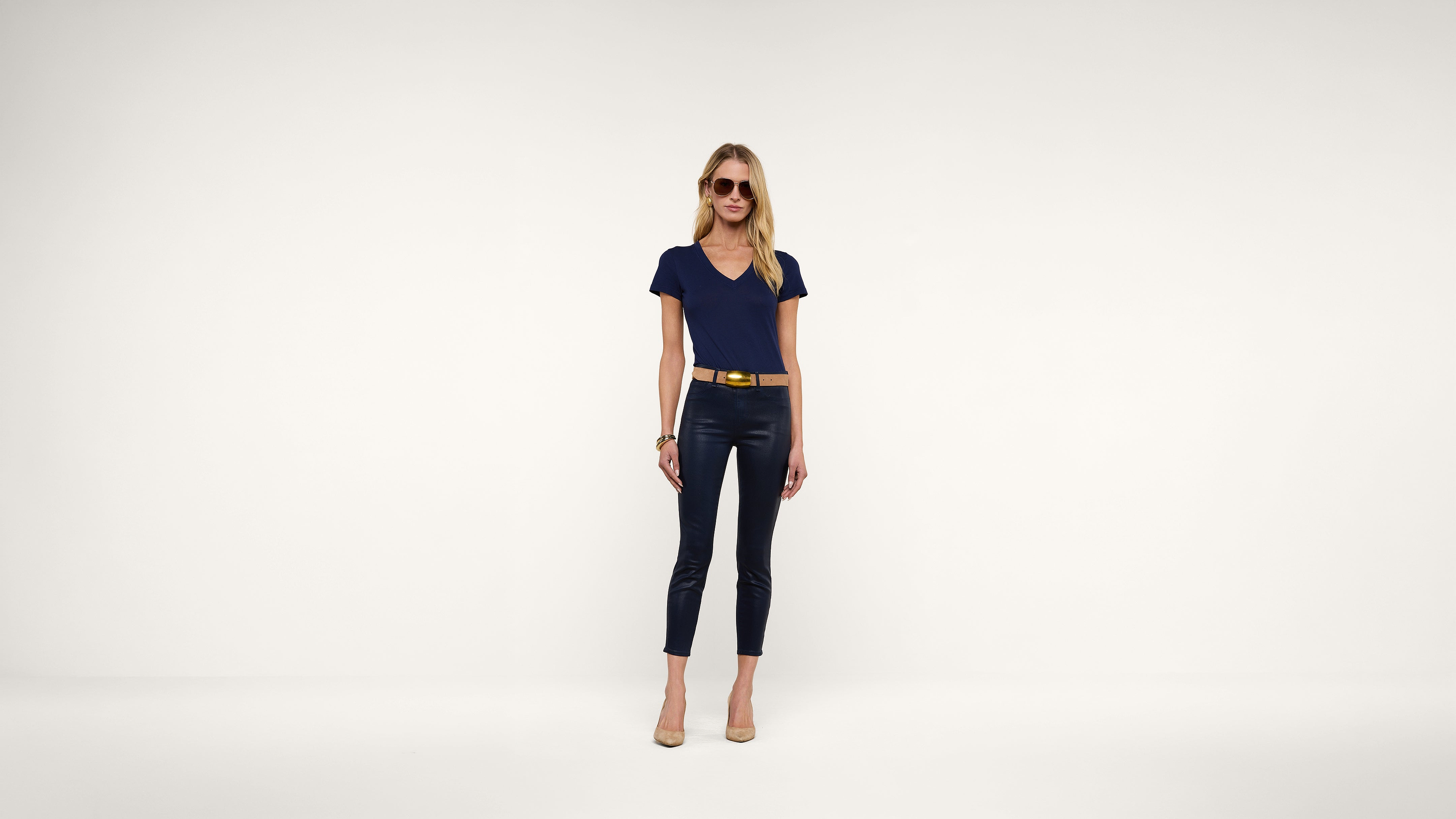 A woman wearing sunglasses, a navy t-shirt, black pants, a yellow belt, and nude heels stands against a plain white background.