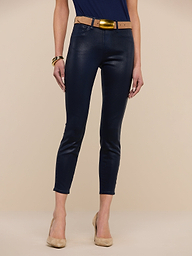 A woman stands wearing the Margot Coated Cropped Skinny Jean, featuring a contoured waistband and tapered leg in navy blue, paired with a tan belt, tan high heels, and a blue top. Her arms are relaxed at her sides with a brown bracelet on her right wrist. The background is plain white.