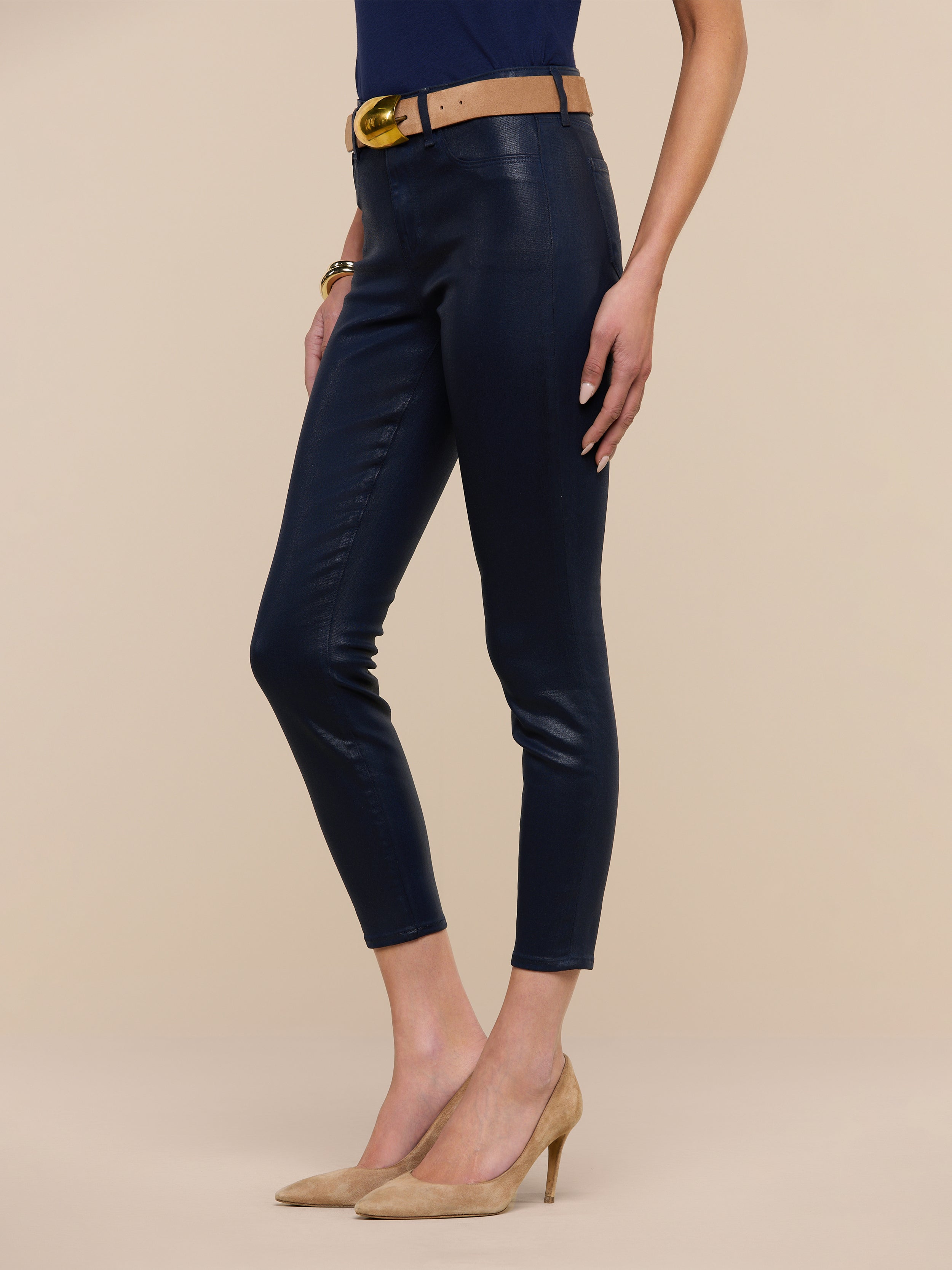 A woman stands against a white background wearing the Margot Coated Cropped Skinny Jean in navy with a tapered leg, styled with a tan belt featuring a gold buckle, a navy top, and tan high heels.