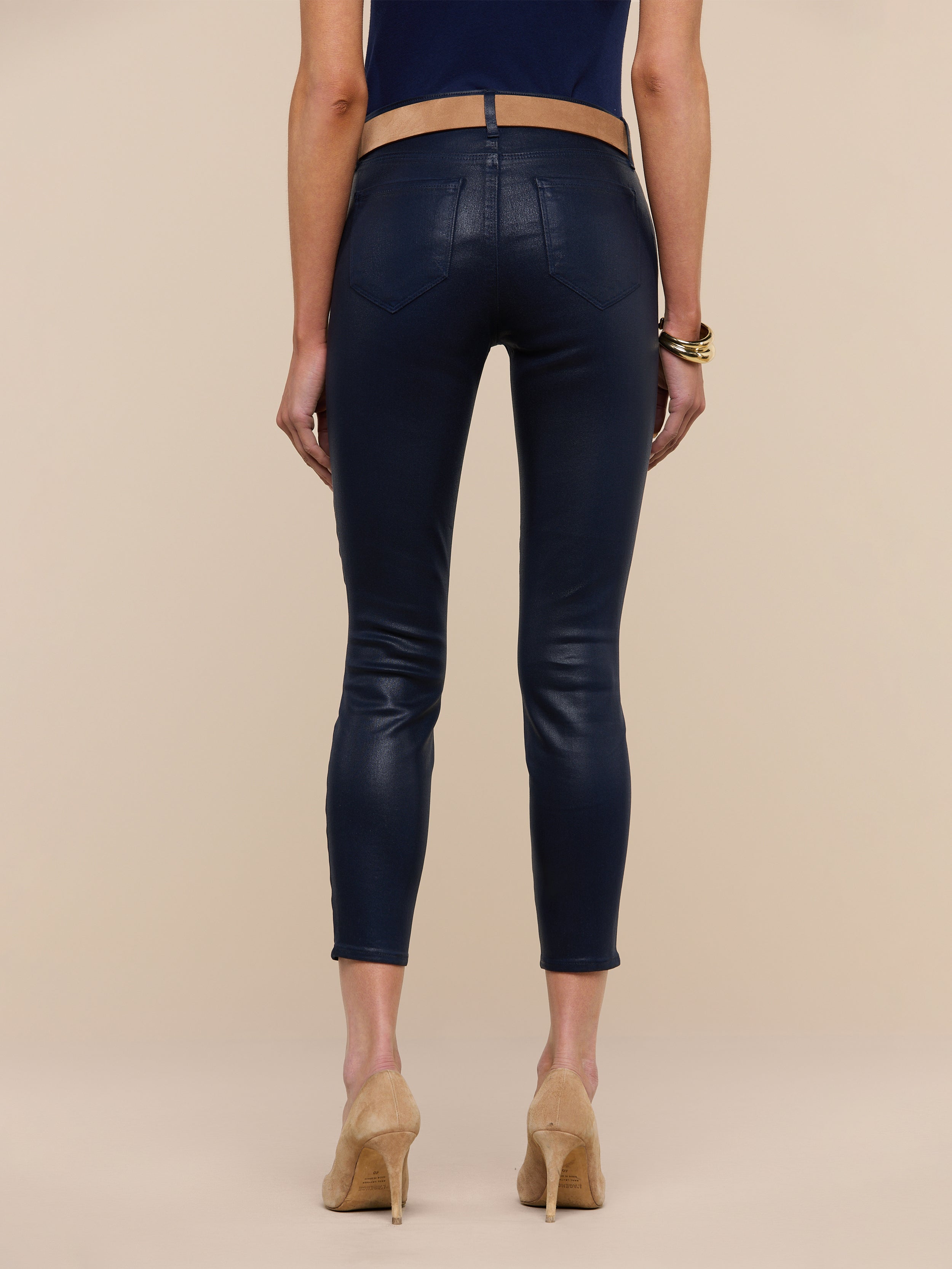 Wearing the Margot Coated Cropped Skinny Jean, which features a contoured waistband and a tapered leg in navy blue, along with a dark top and beige high-heeled shoes, a person stands facing away against a plain white background.