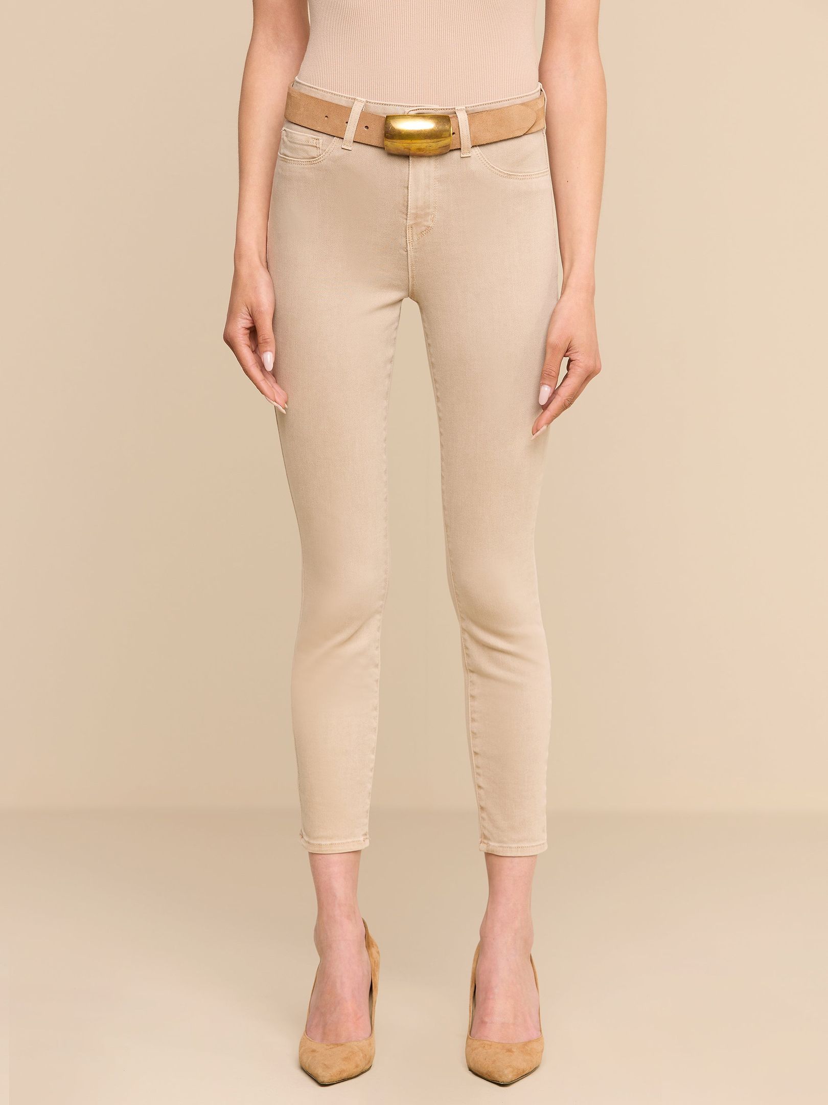 A person stands facing forward, wearing the Margot Cropped Skinny Jean—a beige high-rise skinny jean with a contoured waistband—paired with a beige belt, nude heels, and a beige top against a plain white background.