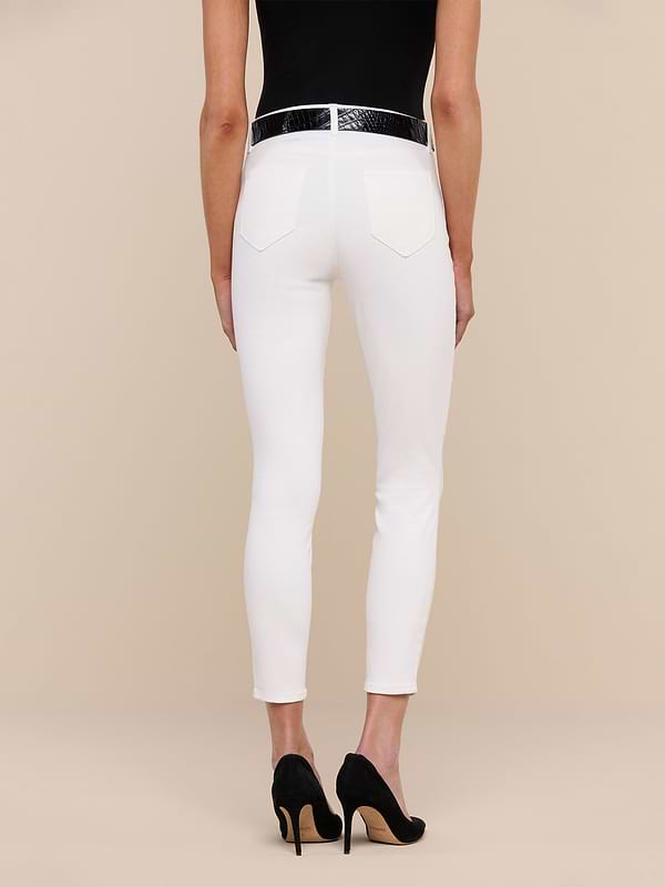 A person stands against a plain white background, shown from the back, wearing a black top and the Margot Cropped Skinny Jean in high-rise skinny white stretch denim with back pockets, paired with a black belt and black high-heeled shoes.