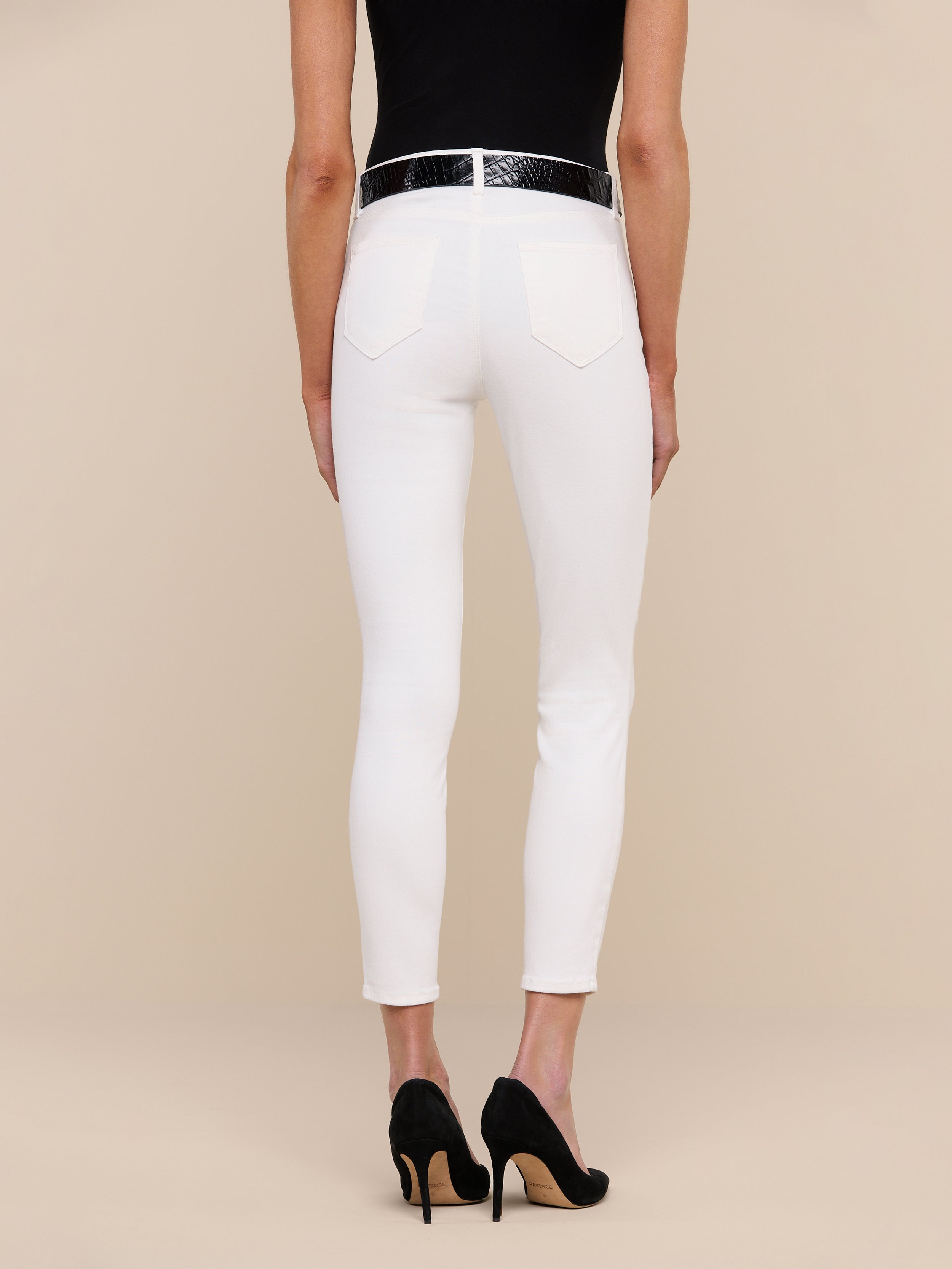 A person stands against a plain white background, shown from the back, wearing a black top and the Margot Cropped Skinny Jean in high-rise skinny white stretch denim with back pockets, paired with a black belt and black high-heeled shoes.