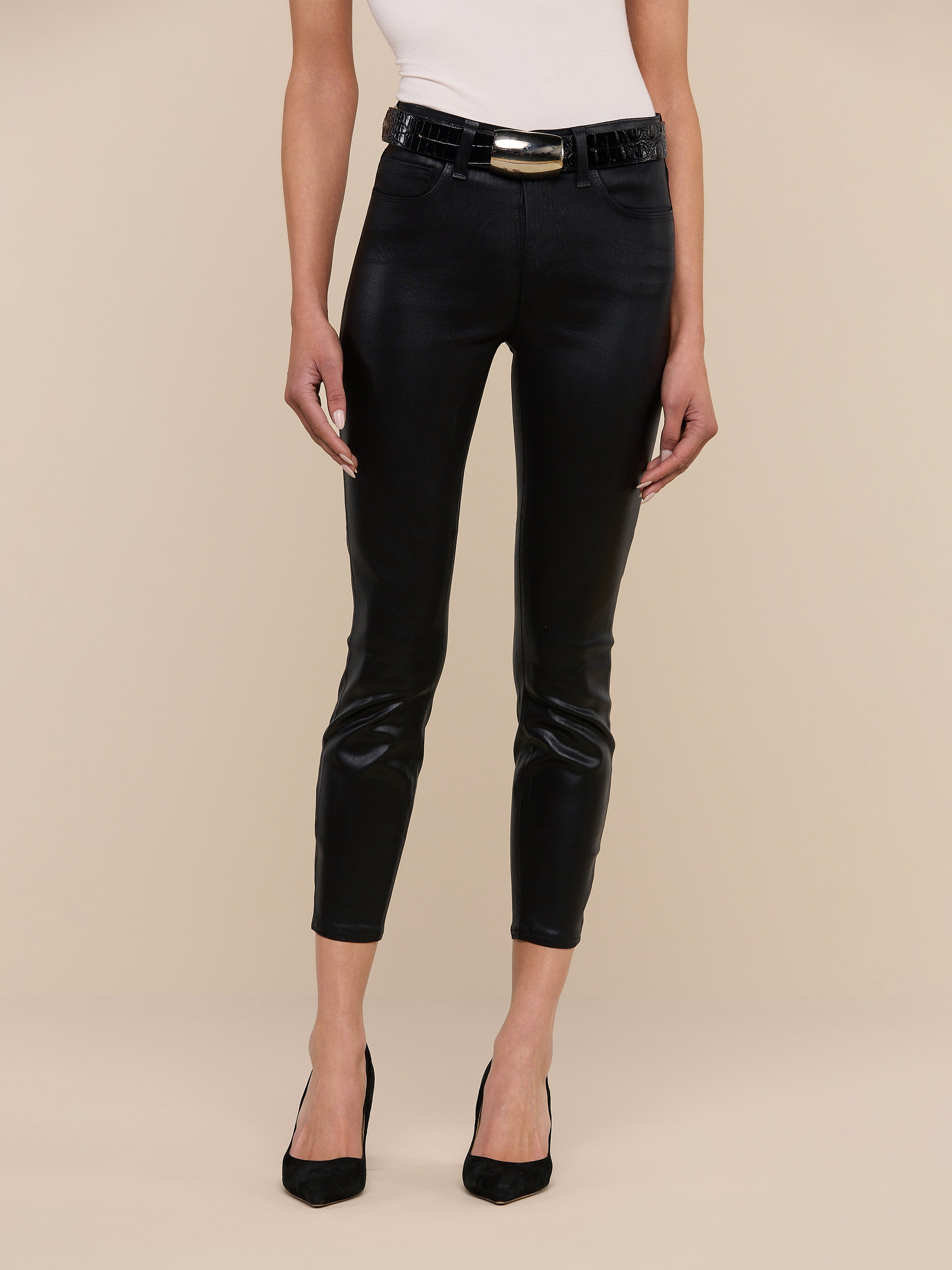 A person wearing a white sleeveless top, black high-heeled shoes, and the Margot Coated Cropped Skinny Jean with a black belt, standing against a plain white background.
