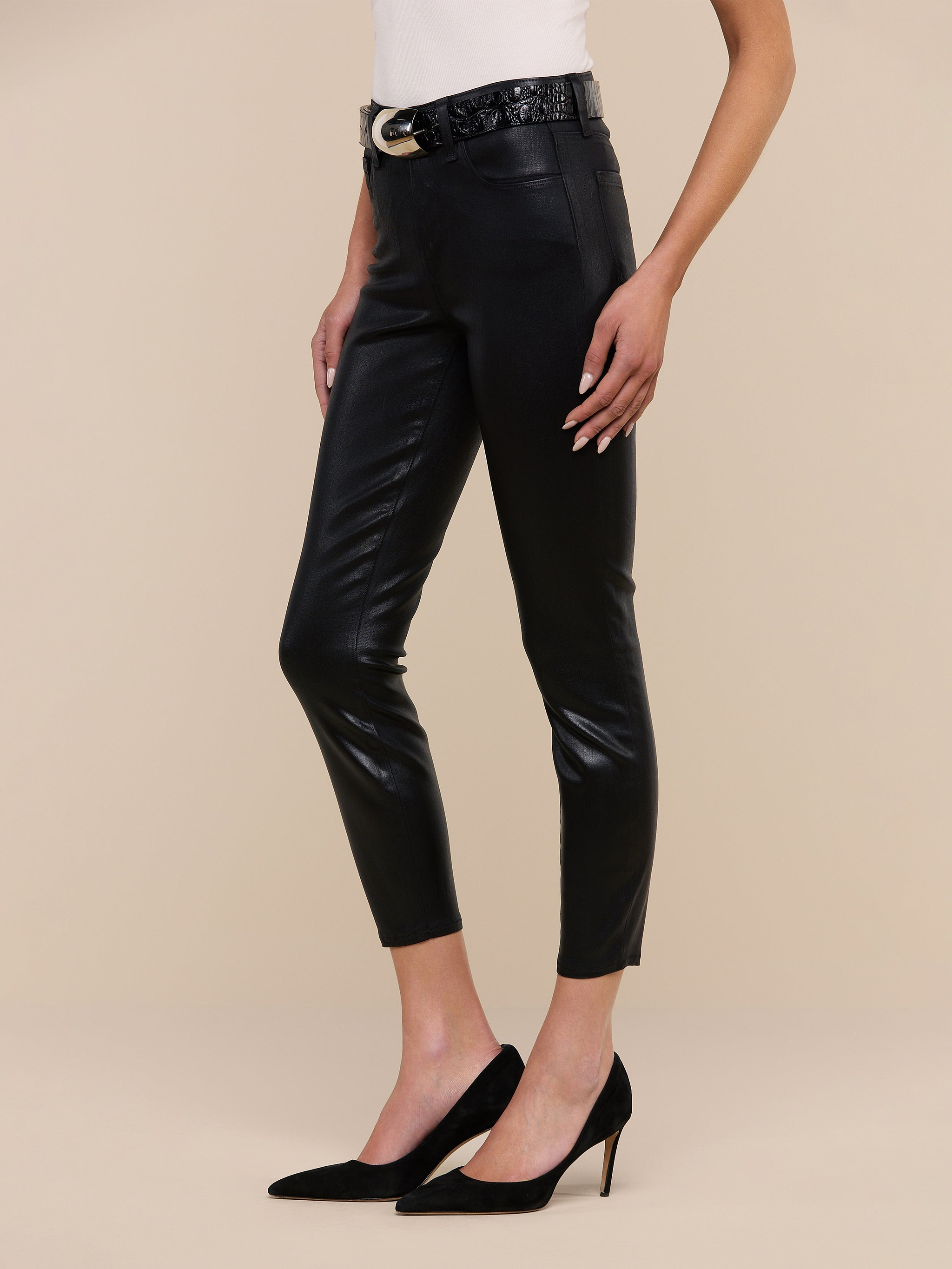 A person wearing the Margot Coated Cropped Skinny Jean, a light top, a black belt with a gold buckle, and black high-heeled shoes poses against a plain white background.