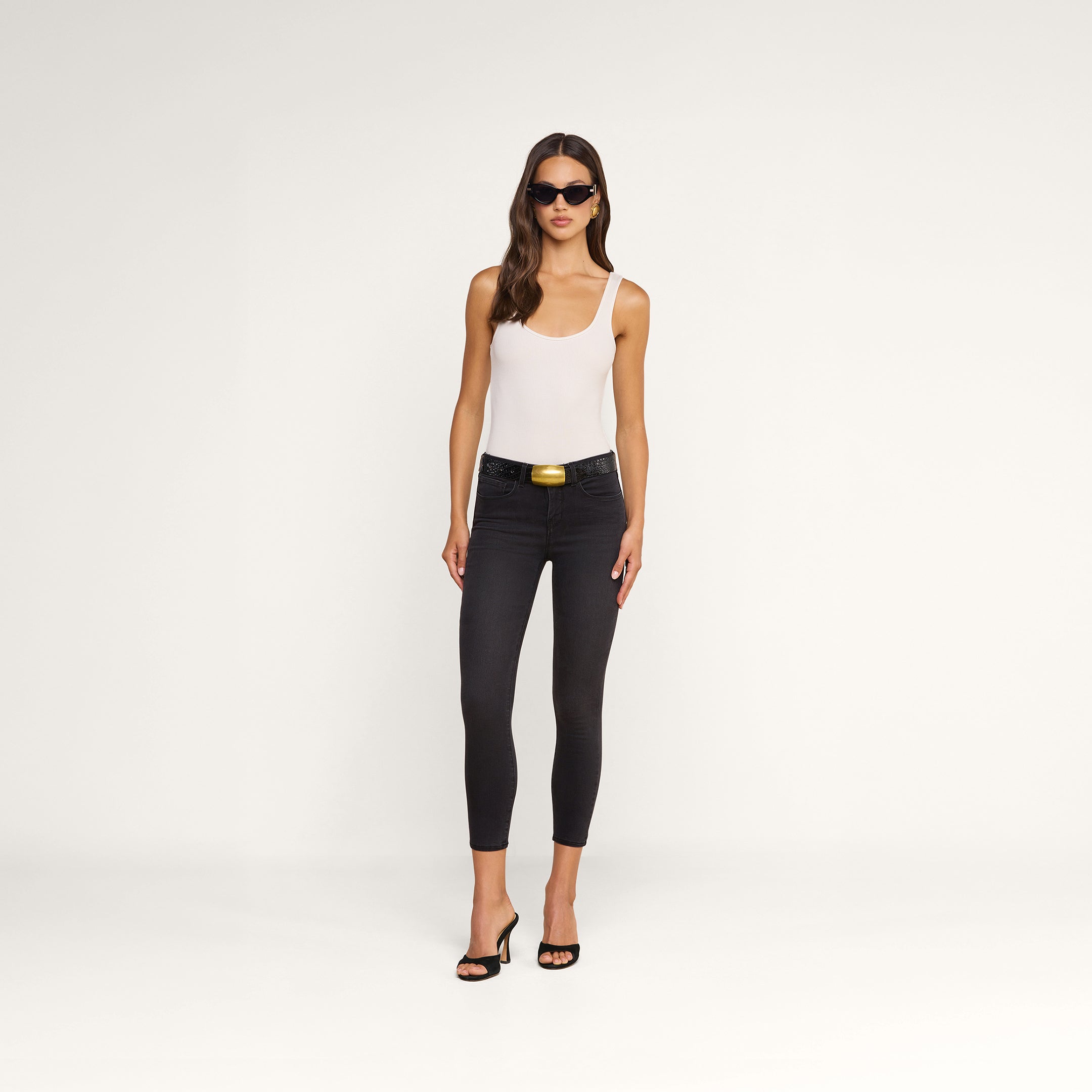 A woman wearing sunglasses, a white sleeveless top, the Margot Cropped Skinny Jean—black high-rise skinny jeans made from stretch denim with a contoured waistband for a flattering fit—a black belt with a gold buckle, and black heels stands against a plain white background.