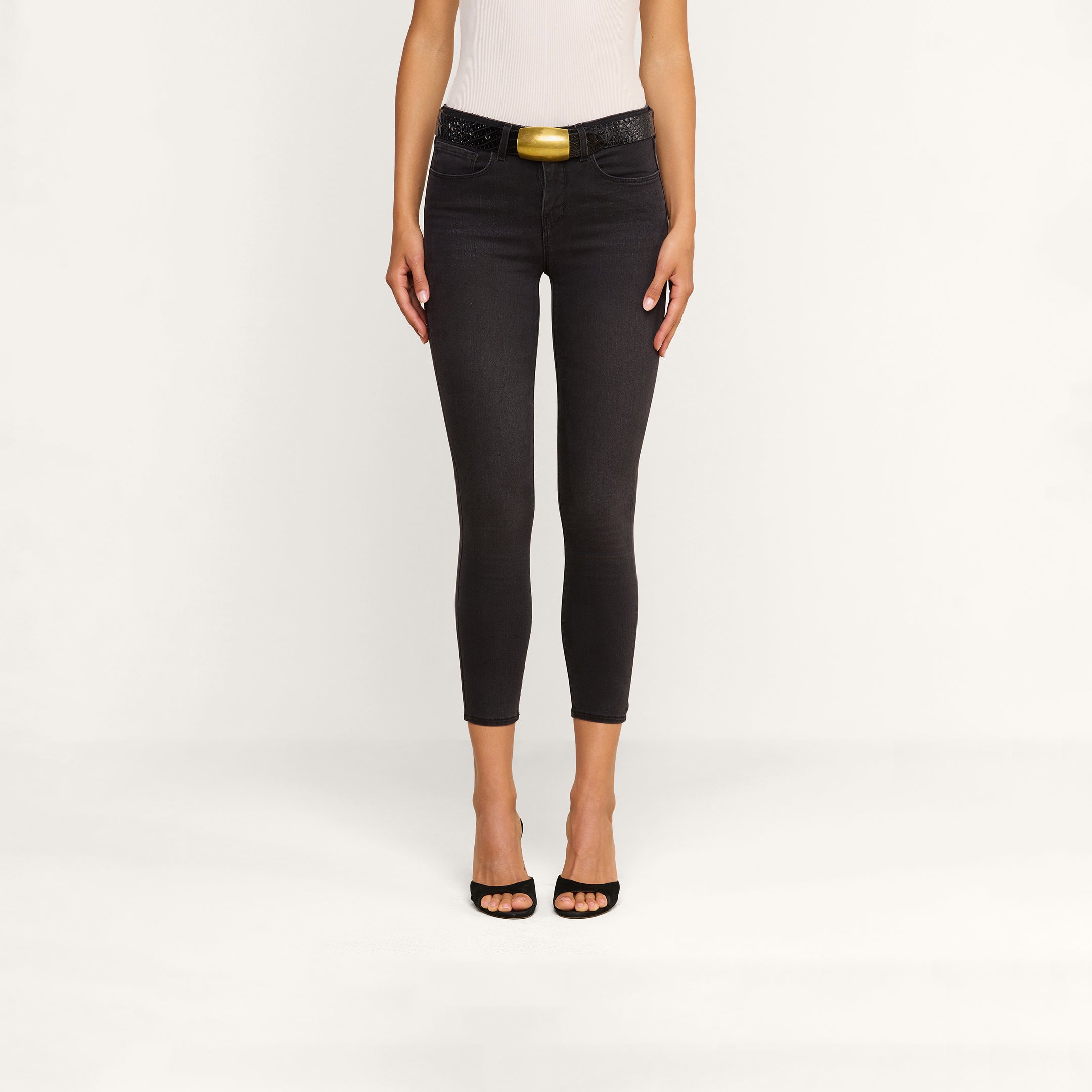 A woman stands against a plain background wearing the Margot Cropped Skinny Jean in black, featuring a contoured high-rise waistband and styled with a belt. She pairs the jeans with a white sleeveless top and black open-toe heels. Her arms are by her sides, and her head is not visible in the image.
