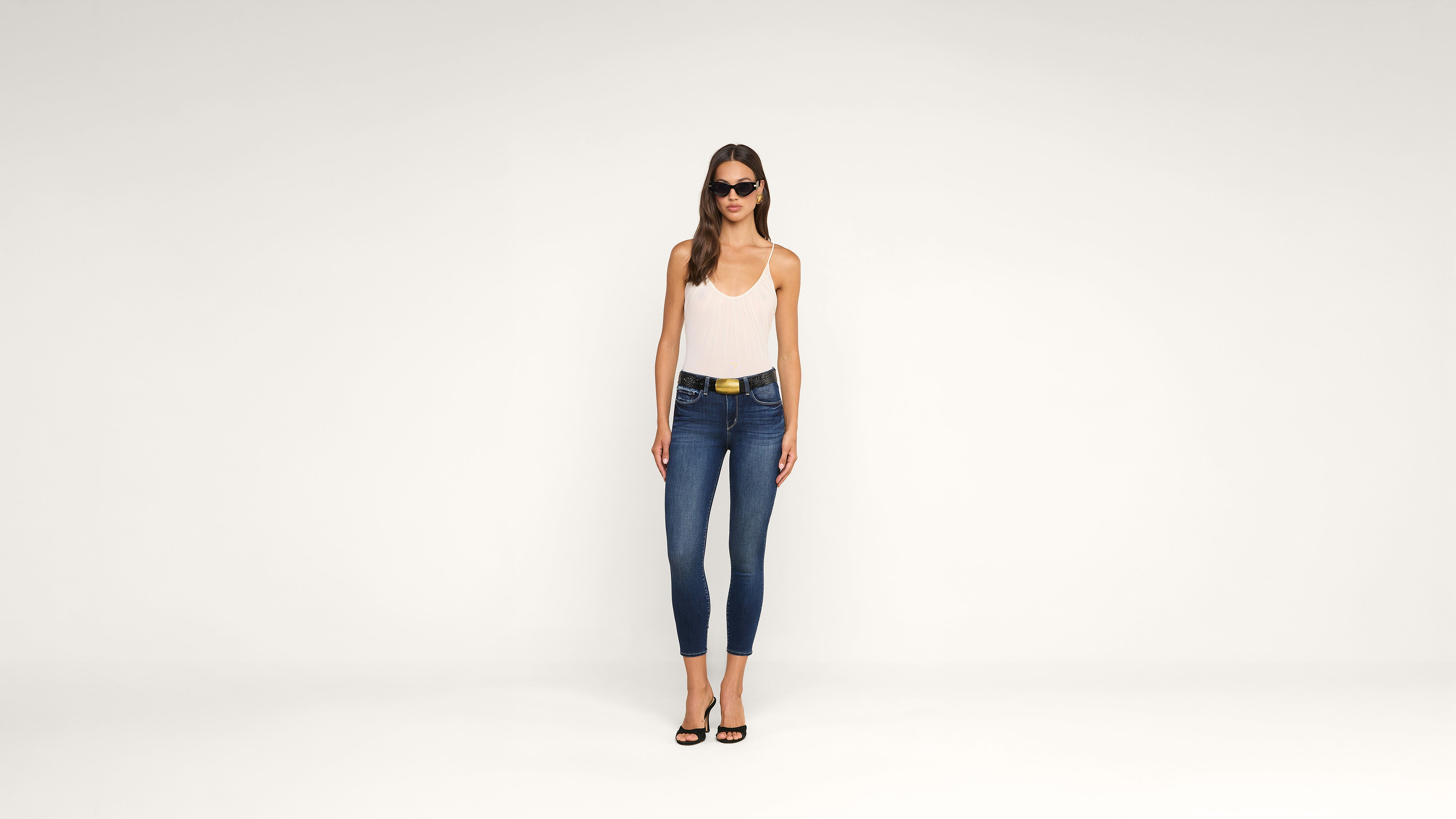 A woman wearing sunglasses, a white sleeveless top, blue skinny jeans, and black heels stands against a plain white background.