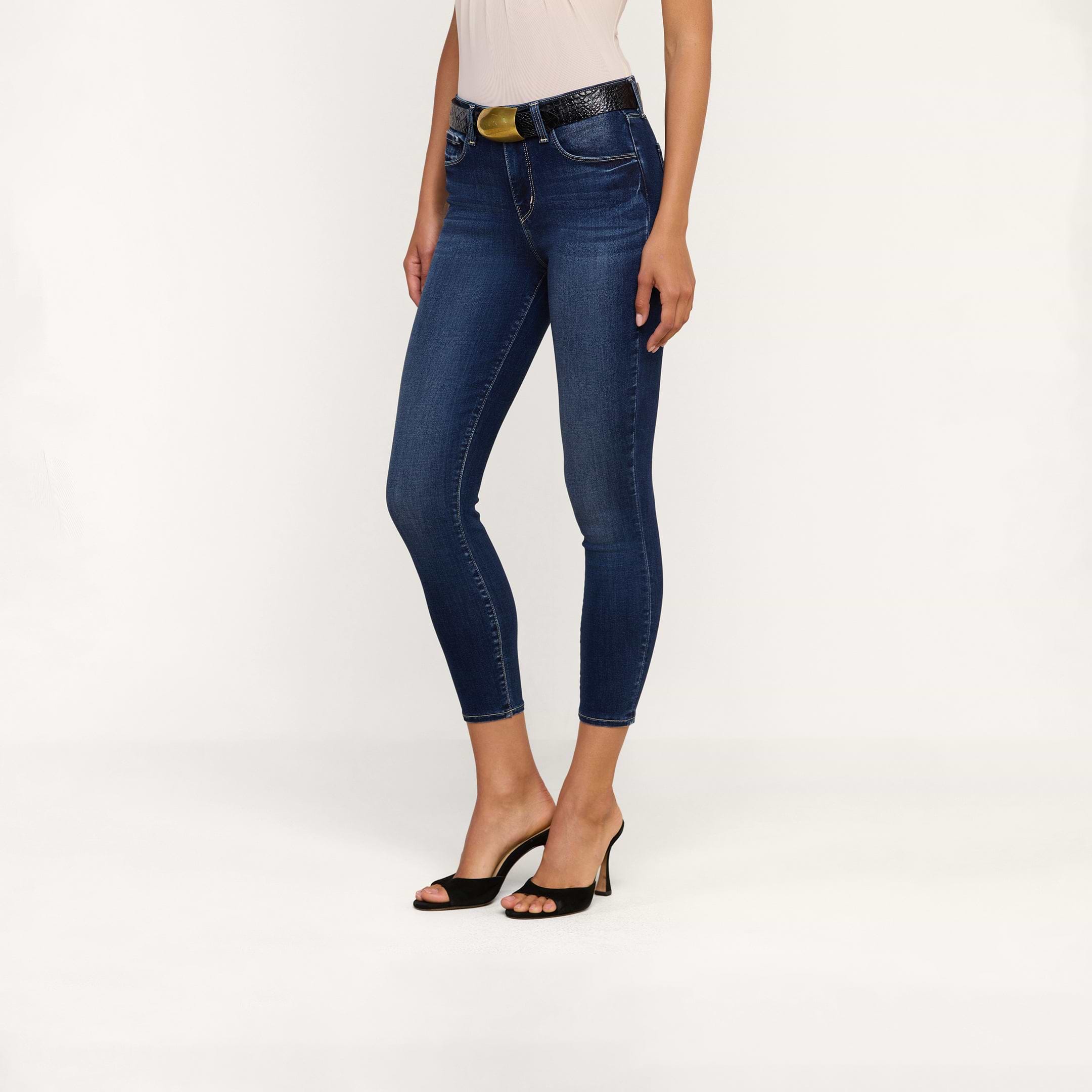 A woman models the Margot Cropped Skinny Jean, crafted from premium stretch denim, styled with a black belt and black open-toe heels. The photo is taken against a plain white background, showing her from shoulders to feet.