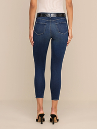 A person stands facing away against a plain white background, wearing a white sleeveless top, a black belt, black heeled sandals, and dark blue Margot Cropped Skinny Jeans crafted from premium stretch denim with back pockets.