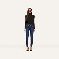 Wearing sunglasses, a black turtleneck sweater, black heels, and the Marguerite Skinny Jean with a belt, a woman stands against a plain, light background.