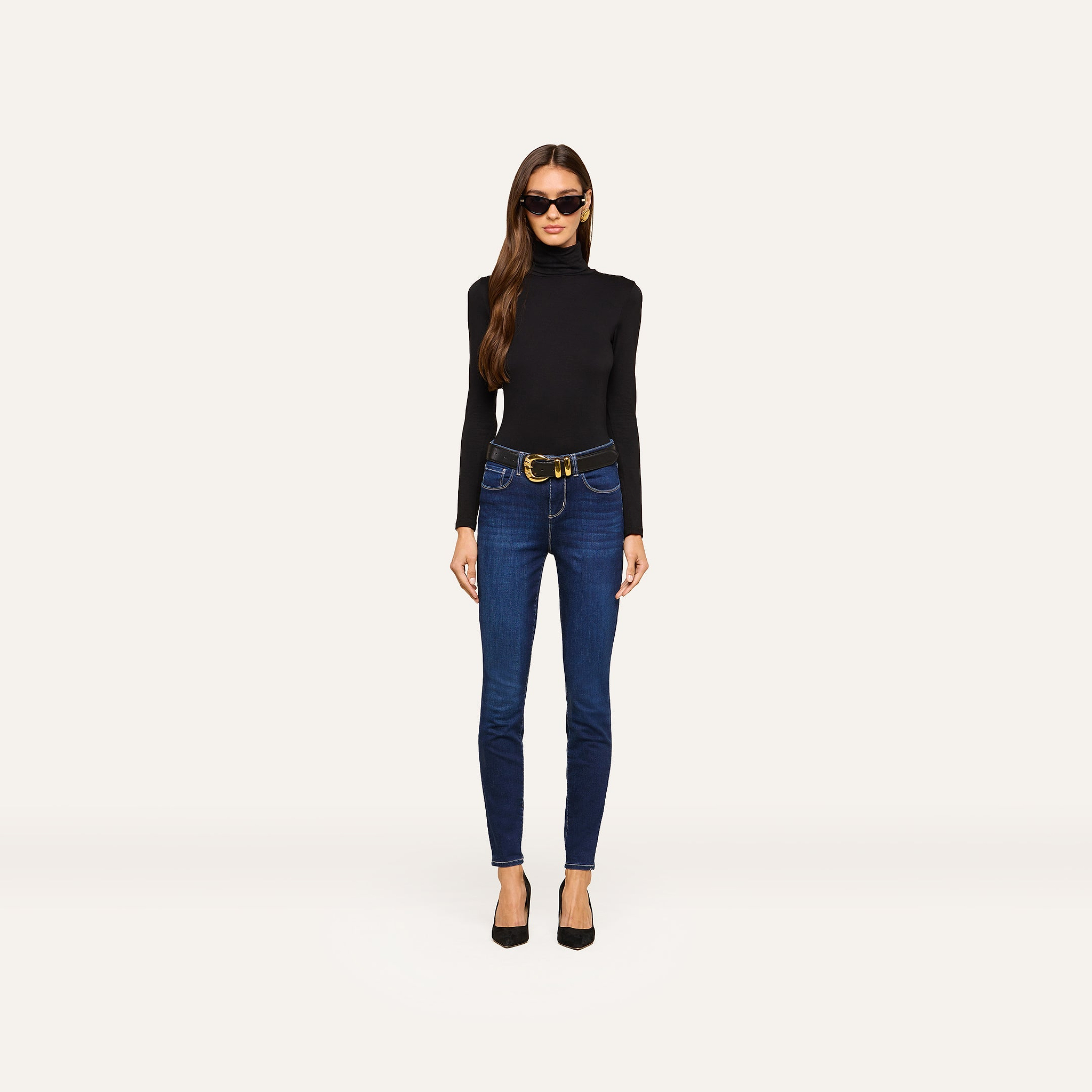 Wearing sunglasses, a black turtleneck sweater, black heels, and the Marguerite Skinny Jean with a belt, a woman stands against a plain, light background.