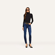 A woman wearing sunglasses, a black long-sleeve turtleneck, a belt, and black high heels stands against a plain light background. She completes her outfit with the Marguerite Skinny Jean—blue high rise skinny jeans crafted from stretch denim.